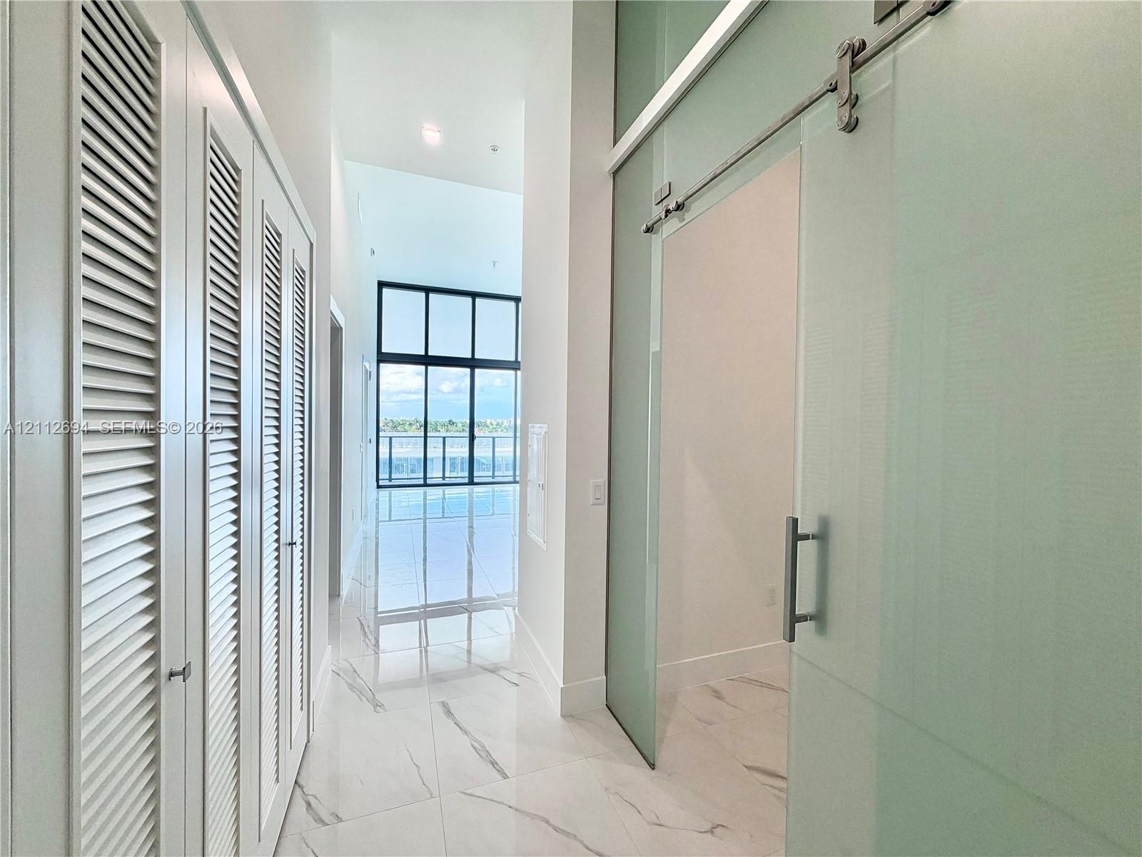 Hallway featuring large-format stone-finish flooring, floor-to-ceiling louvered cabinetry, and a sliding glass door with a polished metal track system Hallway featuring large-format stone-finish flooring, floor-to-ceiling louvered cabinetry, and a sliding glass door with a polished metal track system