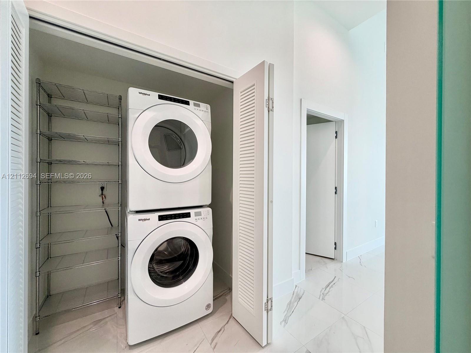 Laundry closet featuring louvered bifold doors, stone-finish tile flooring, and integrated metal shelving Laundry closet featuring louvered bifold doors, stone-finish tile flooring, and integrated metal shelving