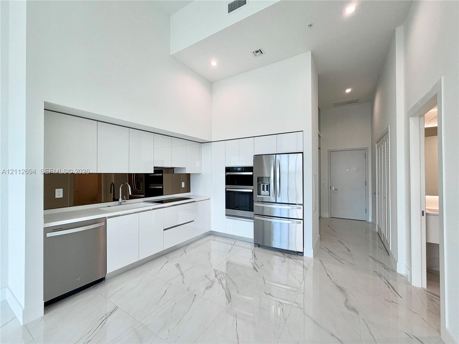 High-gloss white cabinetry, polished stone-finish flooring with veining, stainless steel built-in appliances, a mirrored backsplash, and high ceilings High-gloss white cabinetry, polished stone-finish flooring with veining, stainless steel built-in appliances, a mirrored backsplash, and high ceilings