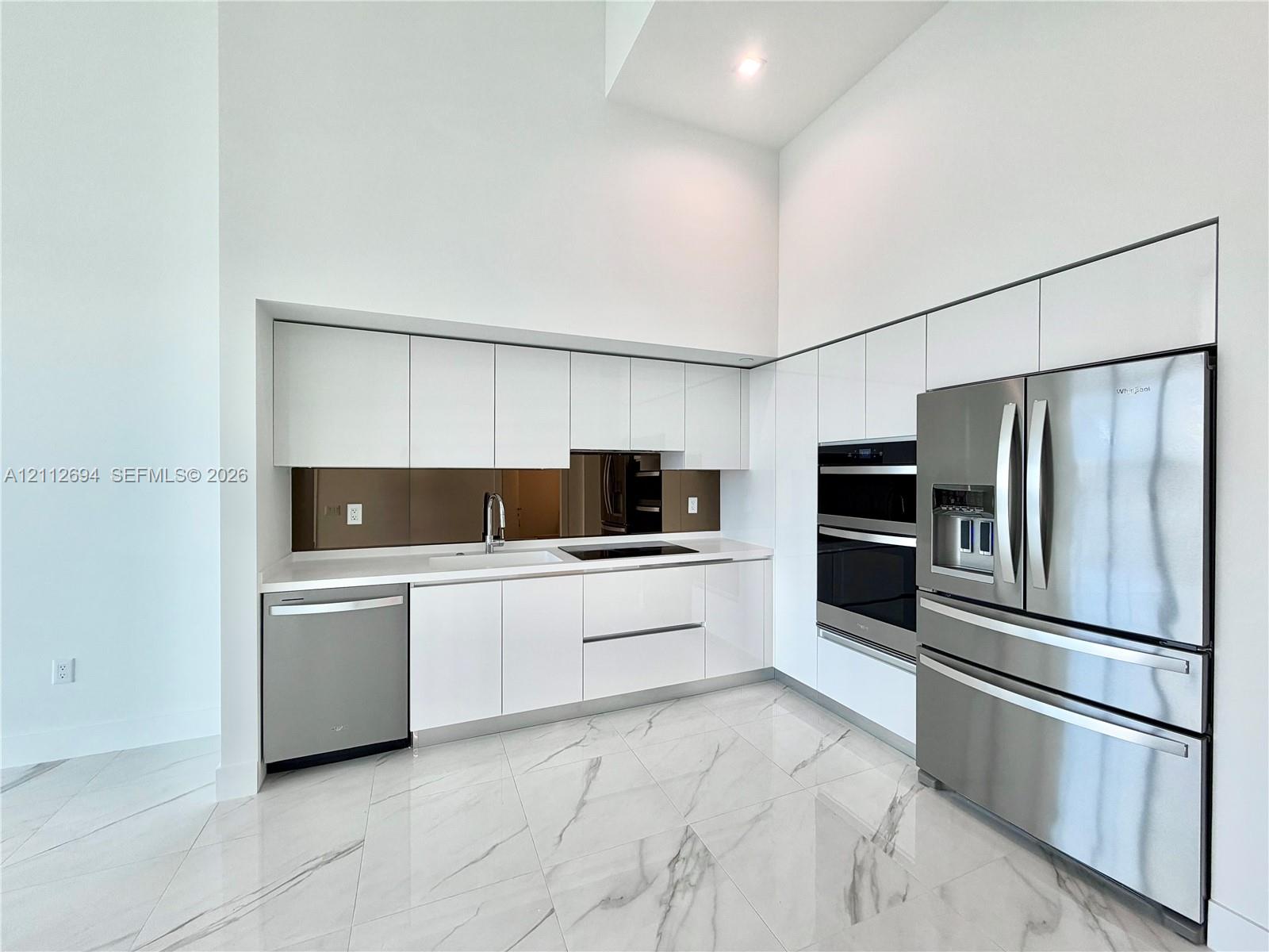 Kitchen featuring high-gloss white cabinetry, stainless steel appliances, built-in dual wall ovens, solid-surface countertops, and large-format porcelain tile flooring Kitchen featuring high-gloss white cabinetry, stainless steel appliances, built-in dual wall ovens, solid-surface countertops, and large-format porcelain tile flooring