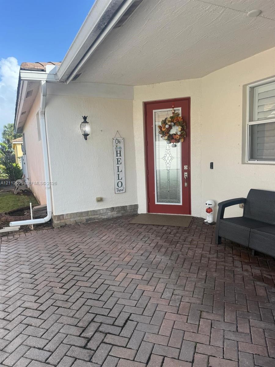 Covered entryway featuring brick-paver flooring in a herringbone pattern, exterior wall-mounted light fixture, and a decorative entry door with an integrated glass panel Covered entryway featuring brick-paver flooring in a herringbone pattern, exterior wall-mounted light fixture, and a decorative entry door with an integrated glass panel