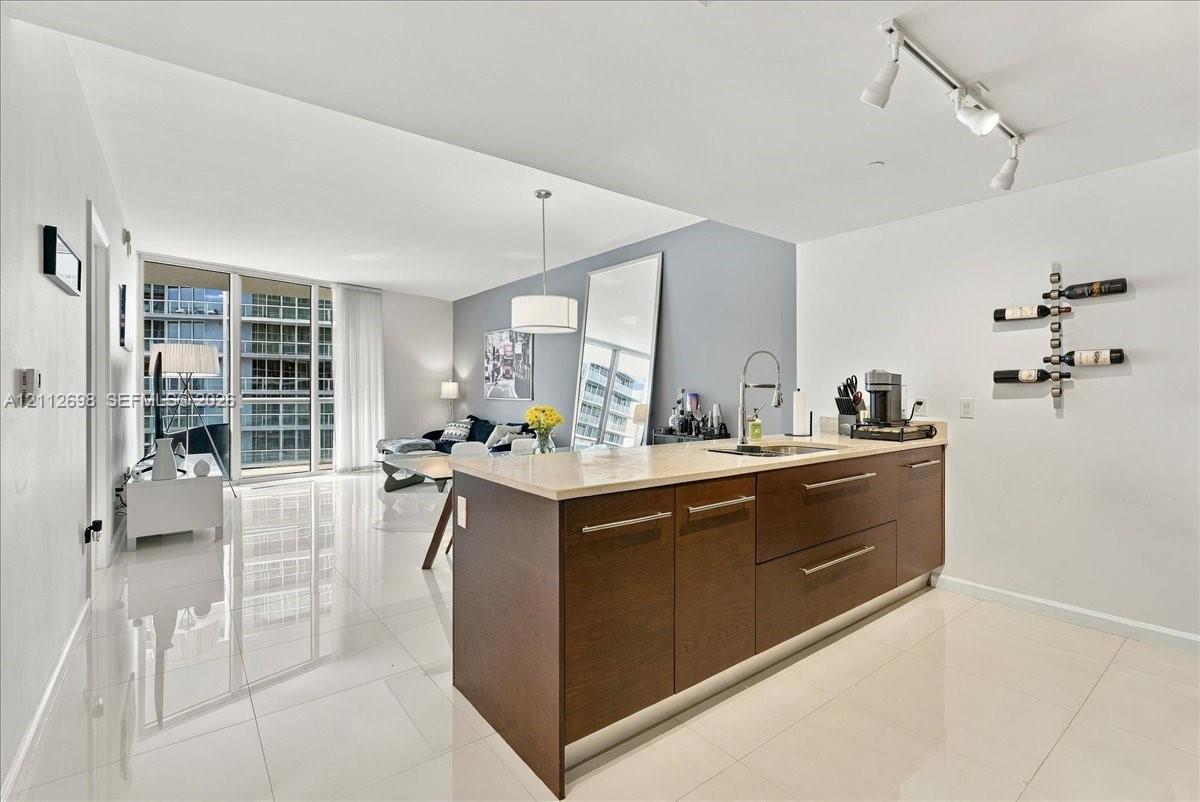 Kitchen island featuring dark wood-finish cabinetry, neutral stone-finish countertops, and an integrated stainless steel undermount sink with a gooseneck faucet, complemented by large-format polished tile flooring