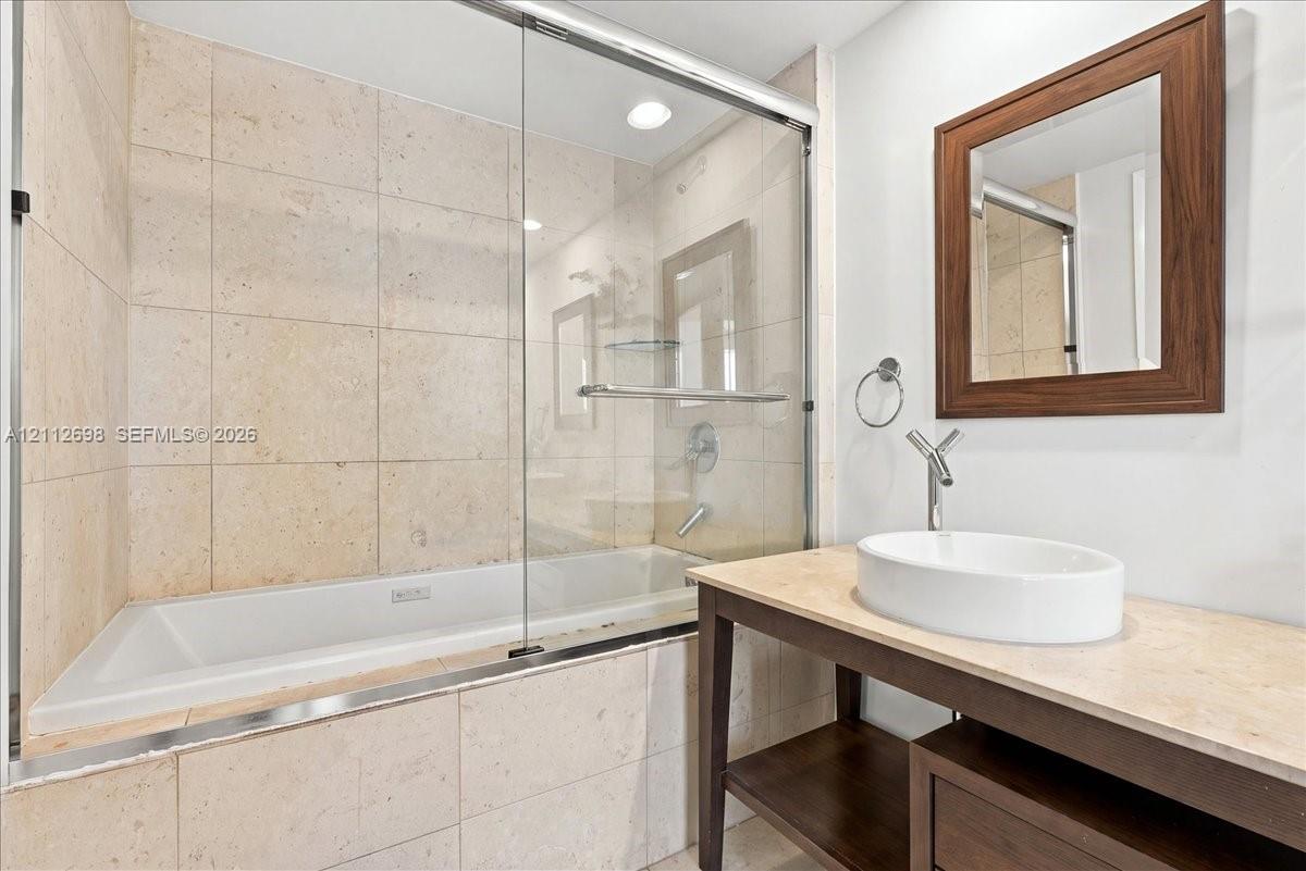 Bathroom featuring a stone-tiled bathtub enclosure with a glass shower door, open-base vanity with a stone countertop, oval vessel sink, and a wood-framed wall mirror