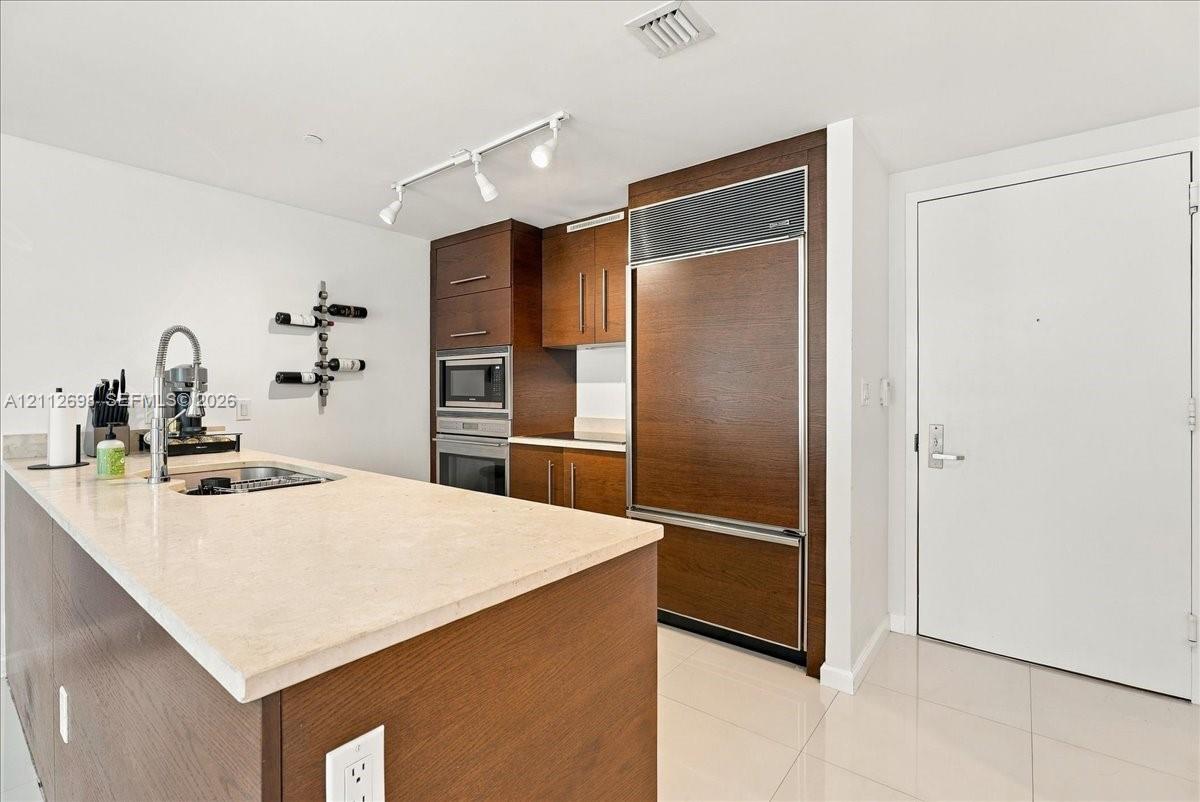 Kitchen featuring wood-finish cabinetry, a paneled refrigerator, integrated wall oven, neutral stone-finish countertops, and track lighting