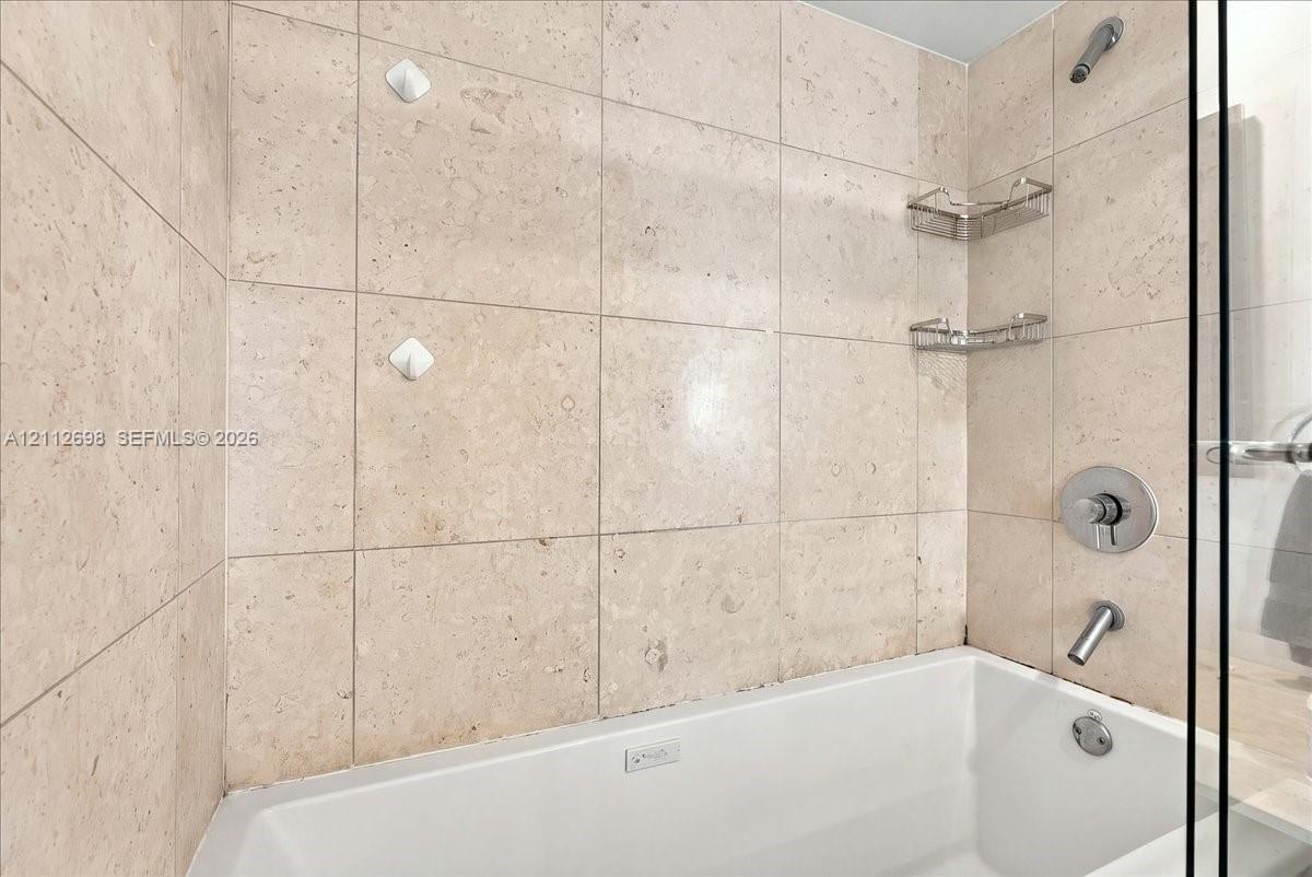 Shower and bathtub combination featuring large-format stone-finish tile surround, polished chrome fixtures, and built-in metal shelving