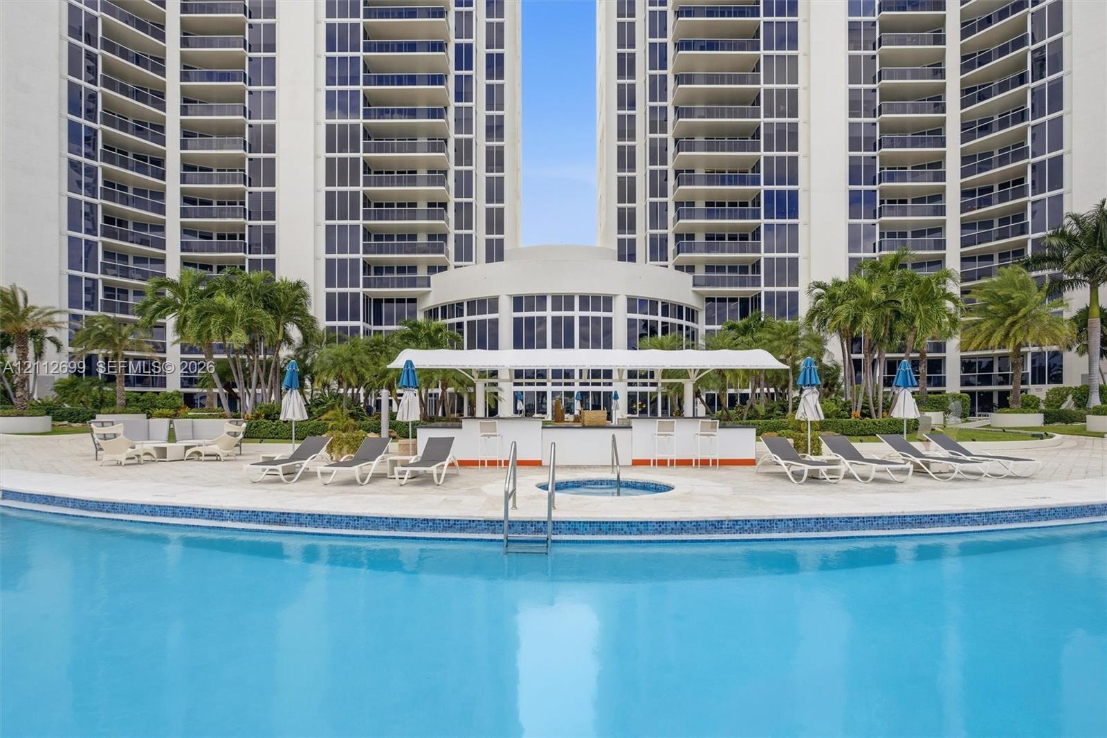 Curved swimming pool with tile trim, integrated spa, poolside bar under a covered canopy, and paved deck featuring mature palm trees