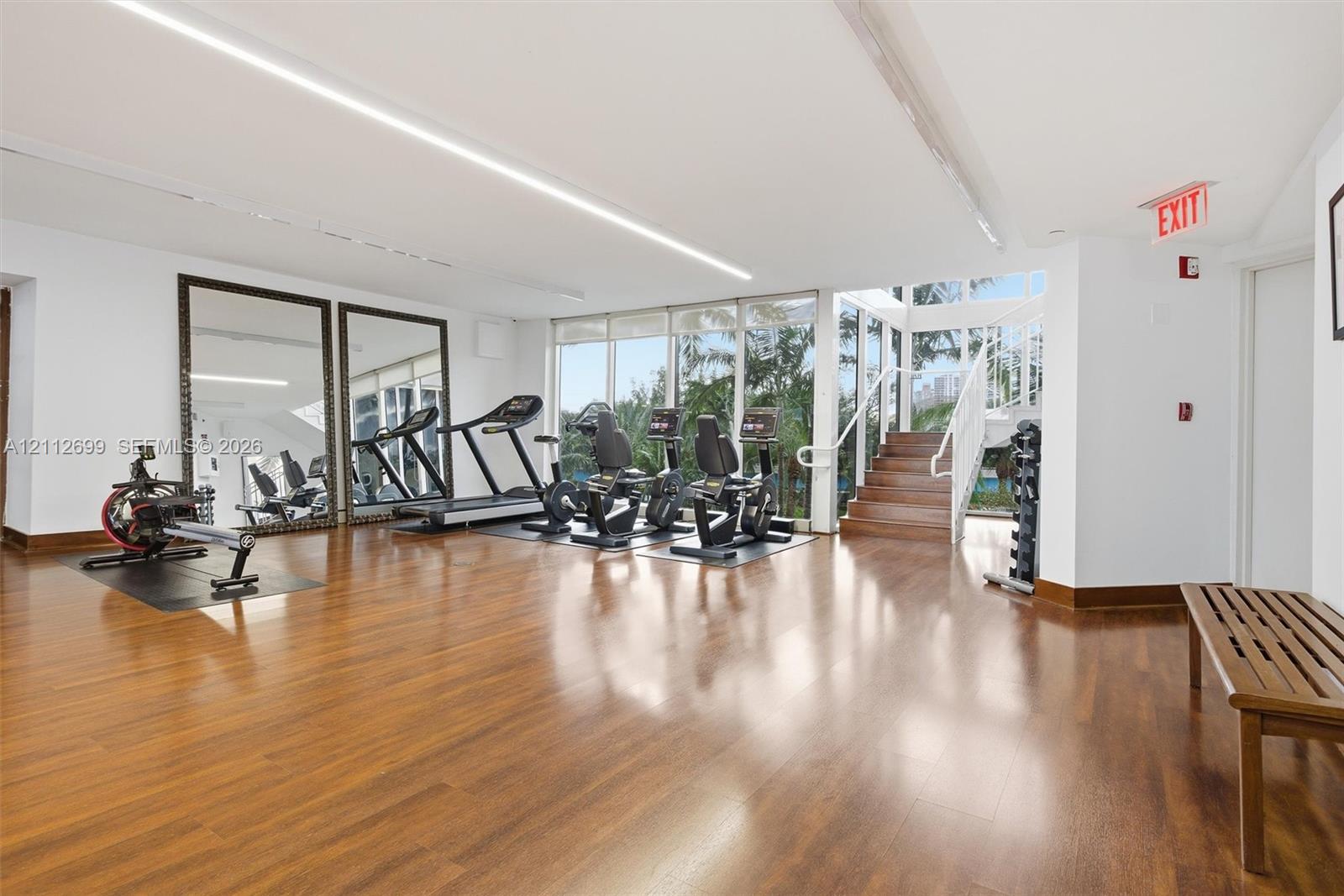 Fitness facility featuring wood-finish flooring, floor-to-ceiling windows, full-wall mirrors, and a wooden staircase with a white metal railing