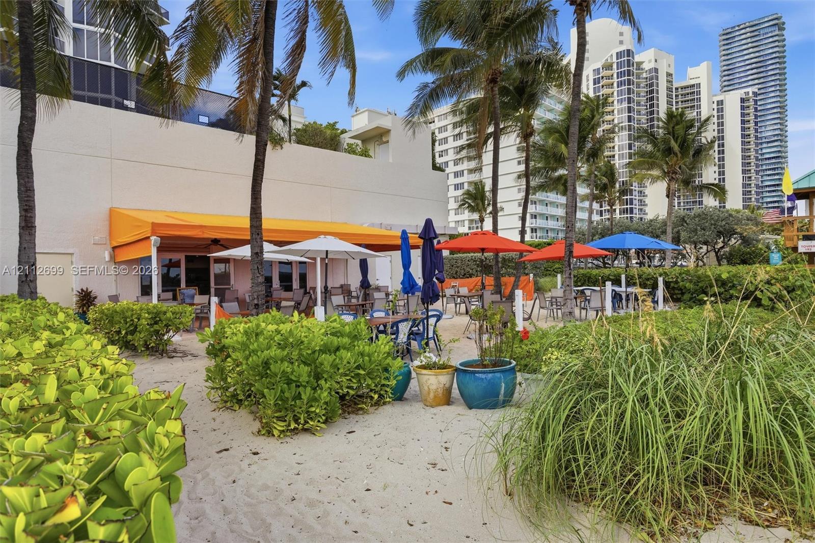 Outdoor patio area featuring a retractable orange awning, sandy ground surface, mature palm trees, and native coastal vegetation