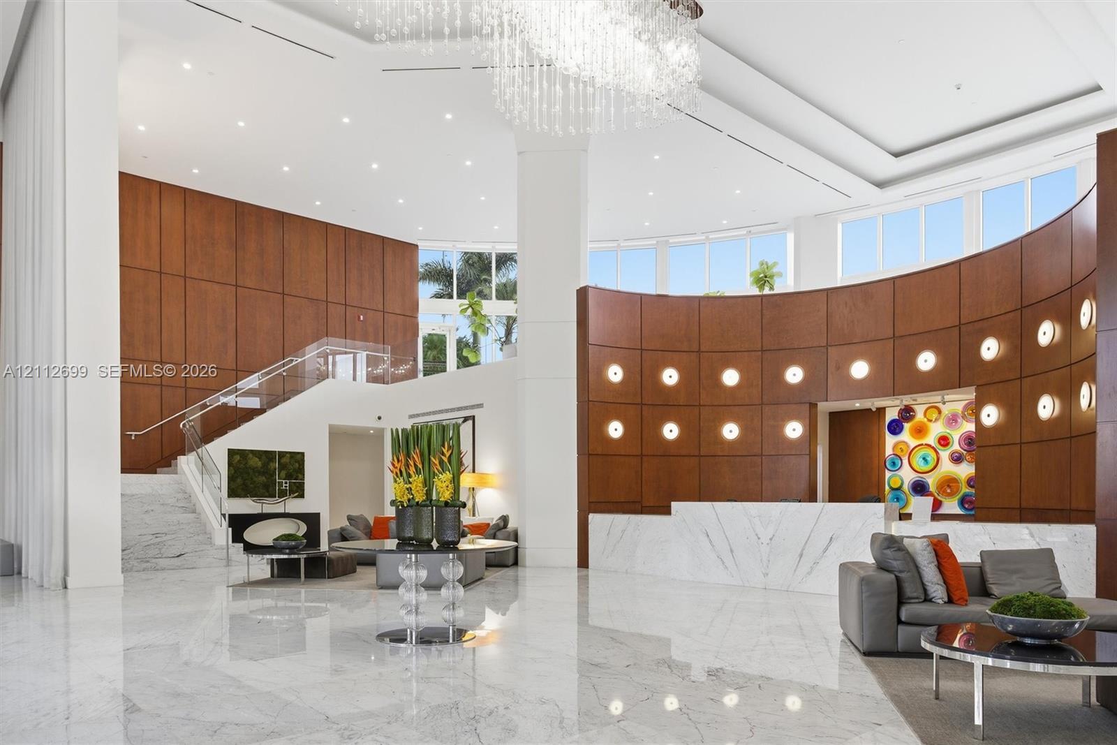 Grand foyer featuring floor-to-ceiling wood paneling, curved architectural walls, polished stone flooring, a multi-tiered crystal chandelier, and a staircase with glass railings