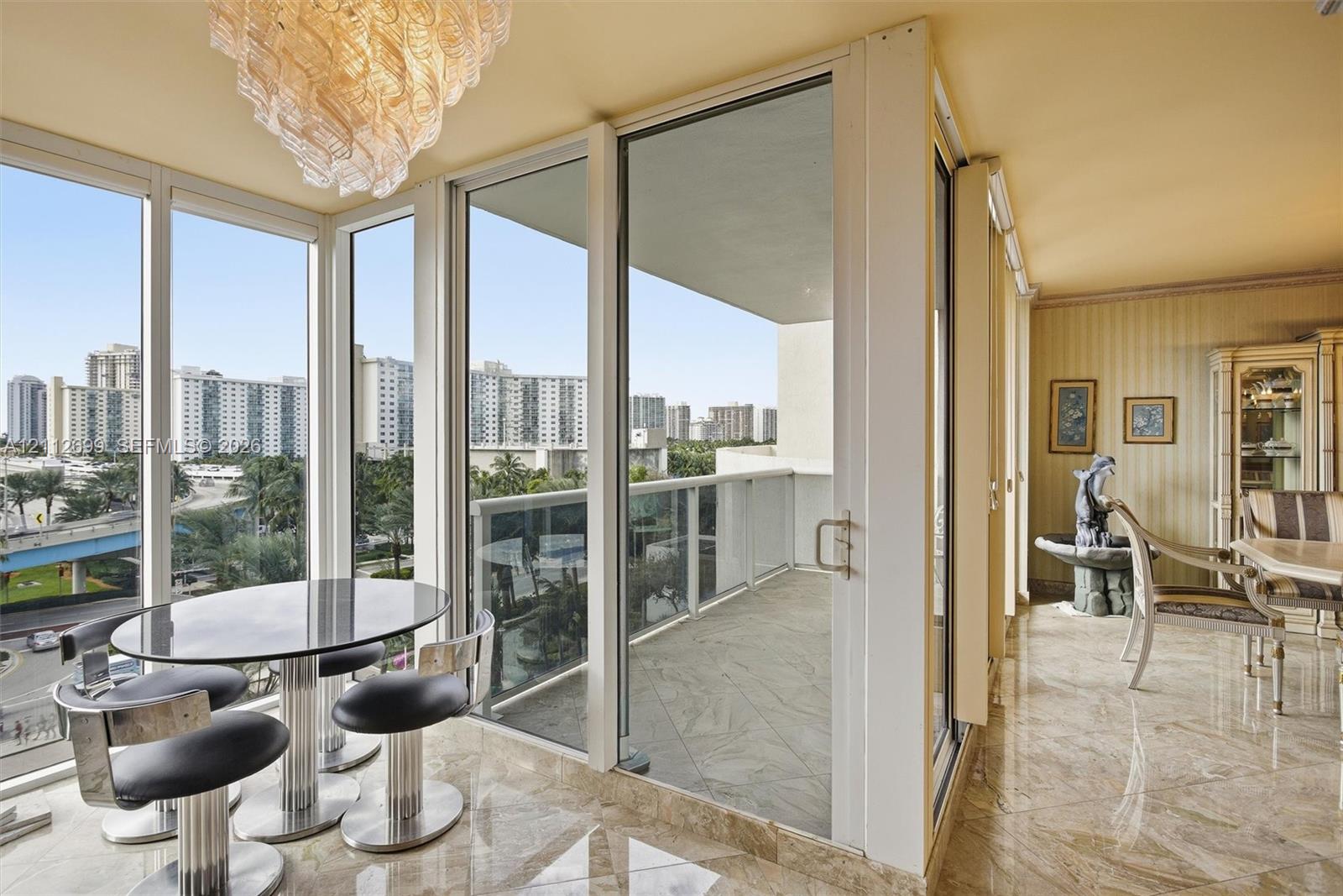 Floor-to-ceiling glass paneling, sliding glass door access to a private balcony, polished stone-finish flooring, and a decorative crystal chandelier