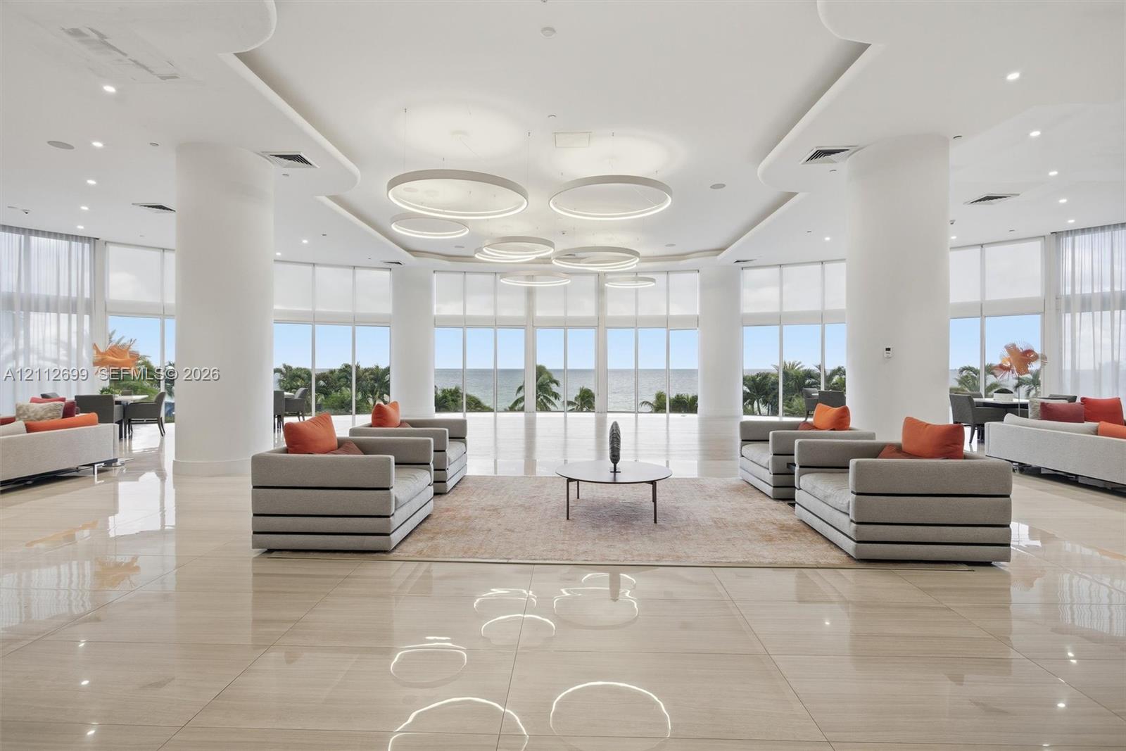 Expansive open-concept space featuring polished stone flooring, floor-to-ceiling windows, sculptural circular pendant lighting, recessed tray ceiling details, and structural support columns