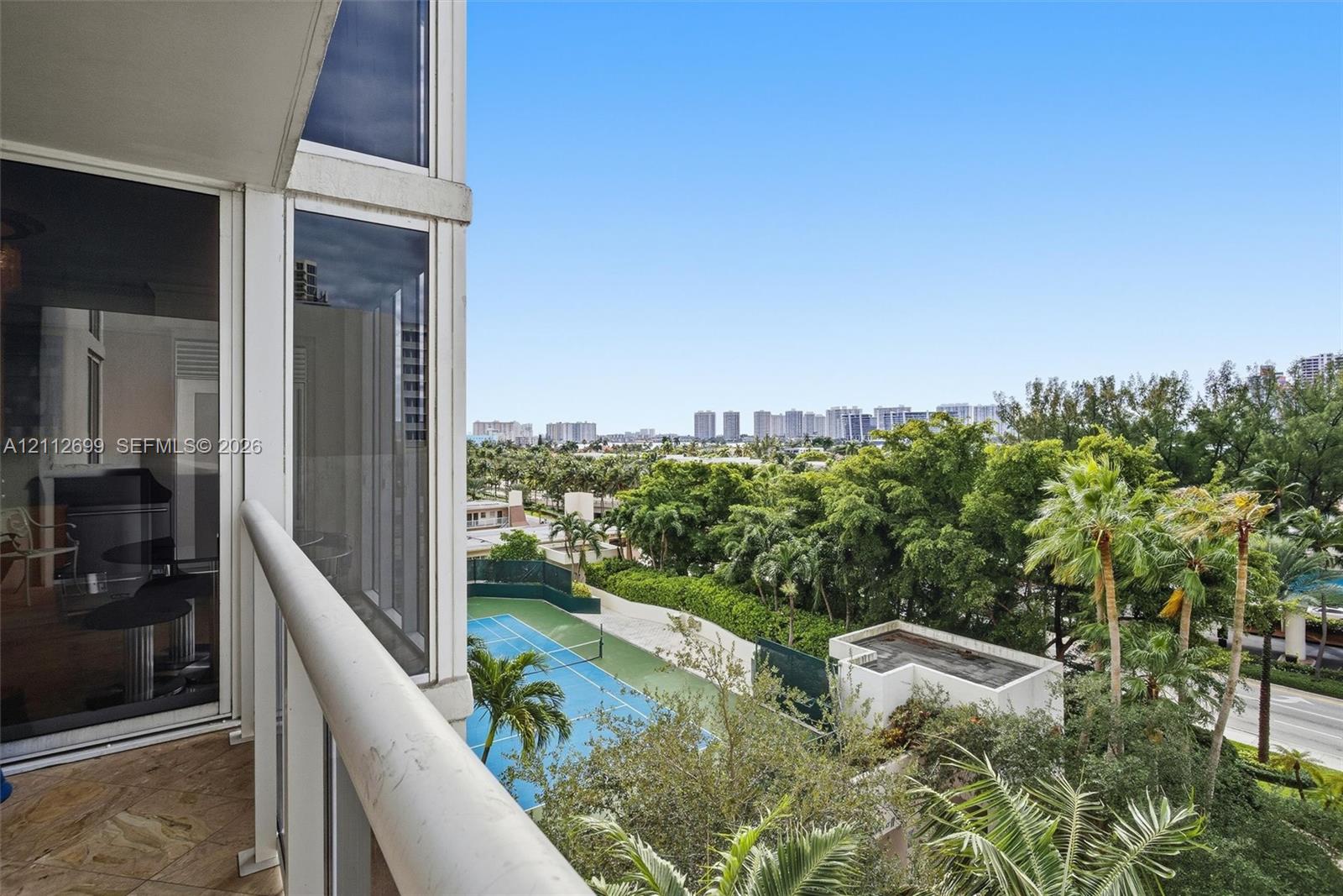 Private balcony featuring stone-finish flooring and a glass-paneled railing, overlooking a tennis court and established palm trees