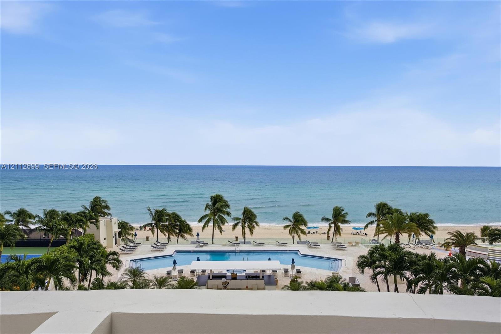 Oceanfront terrace featuring a concrete parapet, overlooking a resort-style swimming pool, lush palm trees, and sand beach