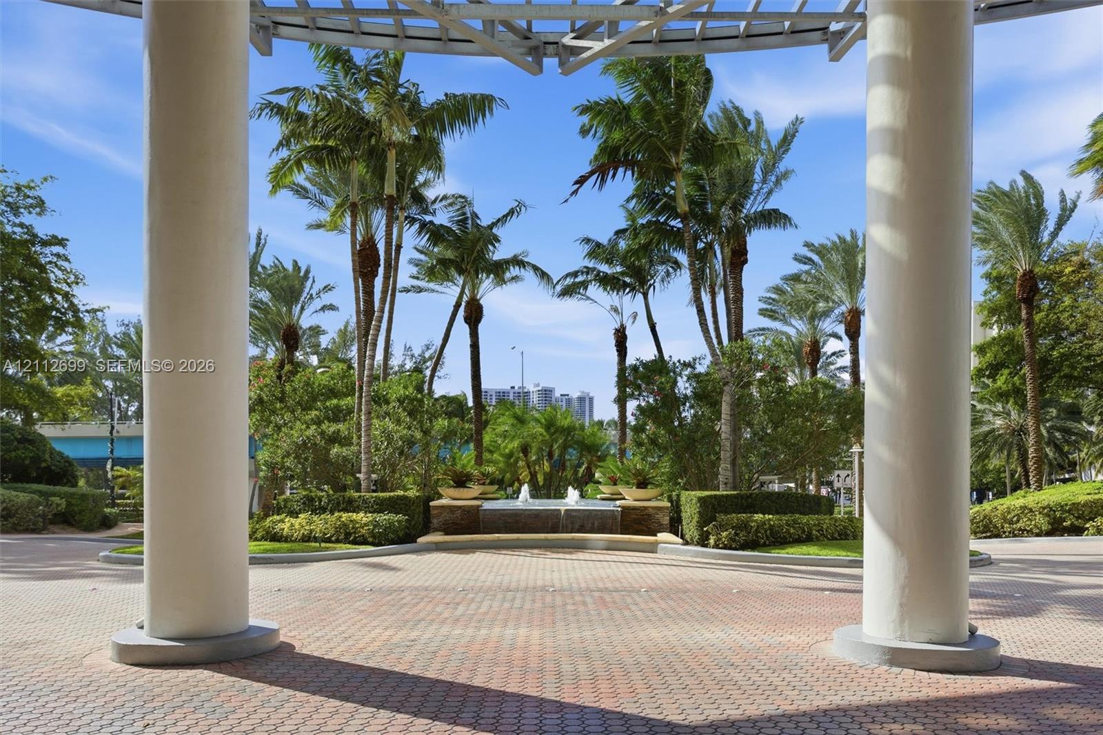 Covered entrance featuring structural columns and a paver stone patio, centered fountain, and mature palm landscaping