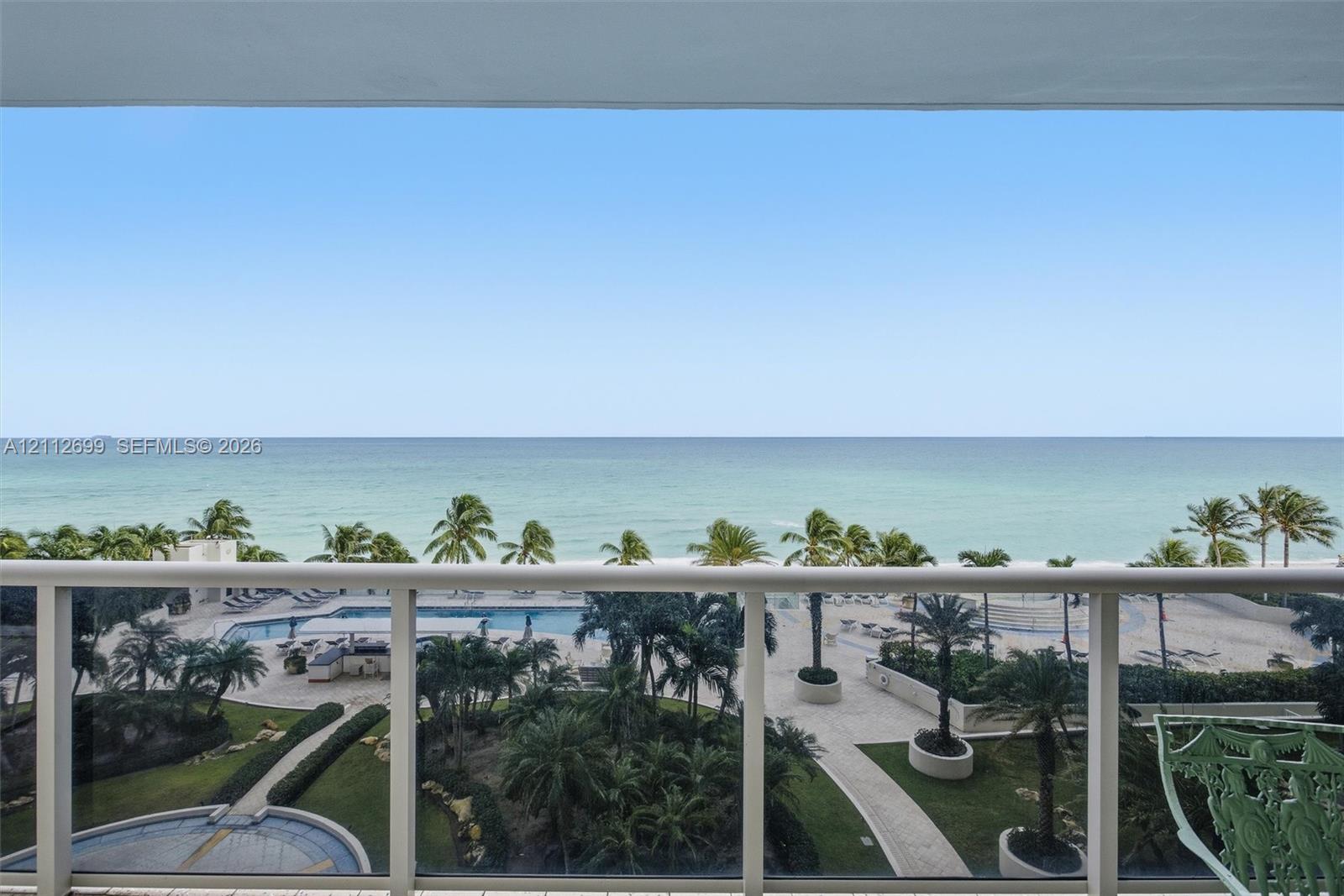 Covered balcony featuring white metal railings, overlooking an oceanfront landscape with a swimming pool, paved walkways, and palm trees