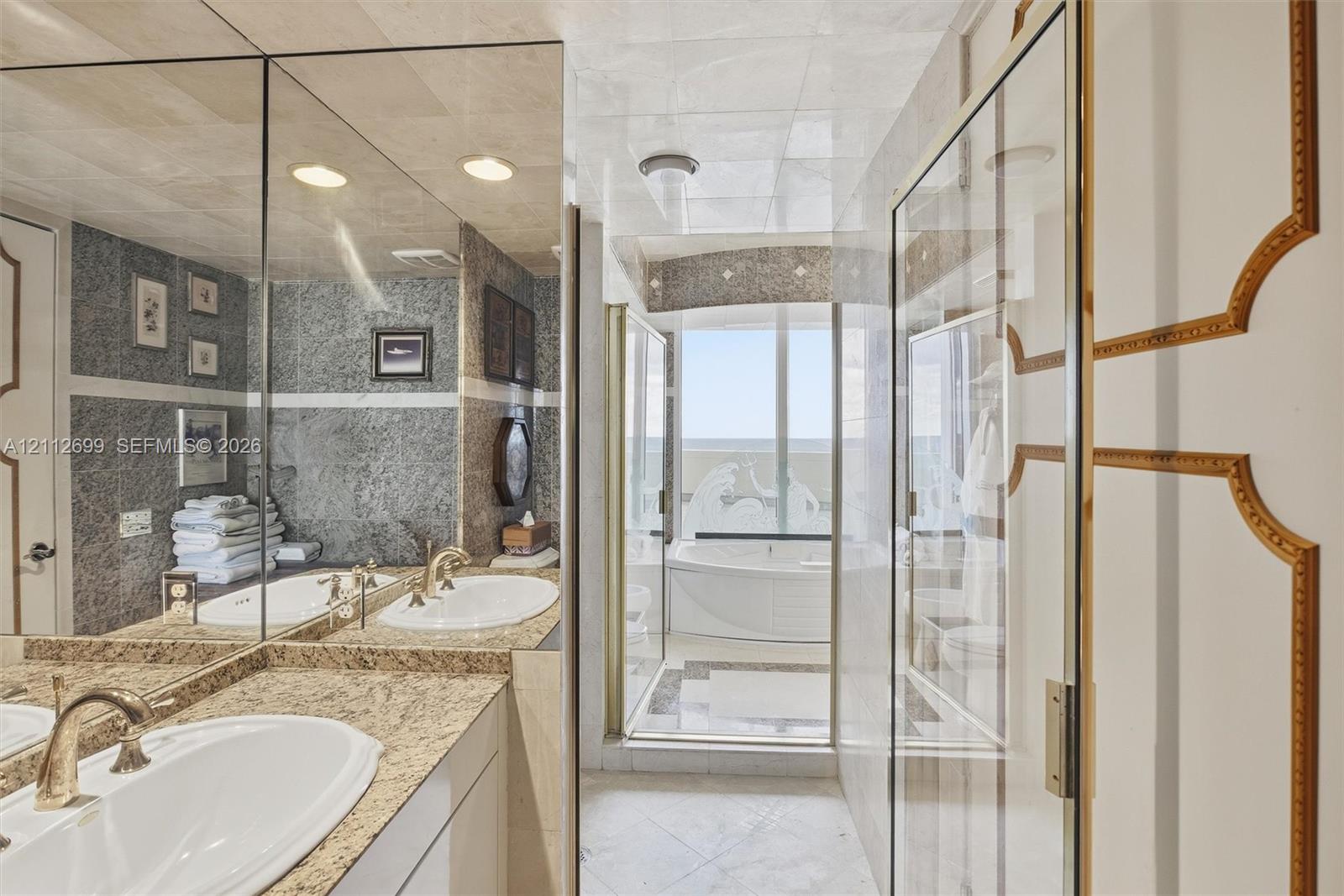 Bathroom featuring a stone-topped dual vanity with integrated sinks, floor-to-ceiling mirrors, tile wall detailing, a jetted soaking tub, and custom paneled doors with gold-tone trim