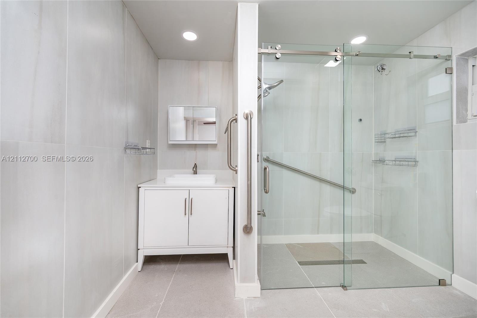 Bathroom featuring large-format wall tile, white vanity cabinet with a rectangular vessel sink, walk-in shower with glass sliding doors, and brushed nickel hardware