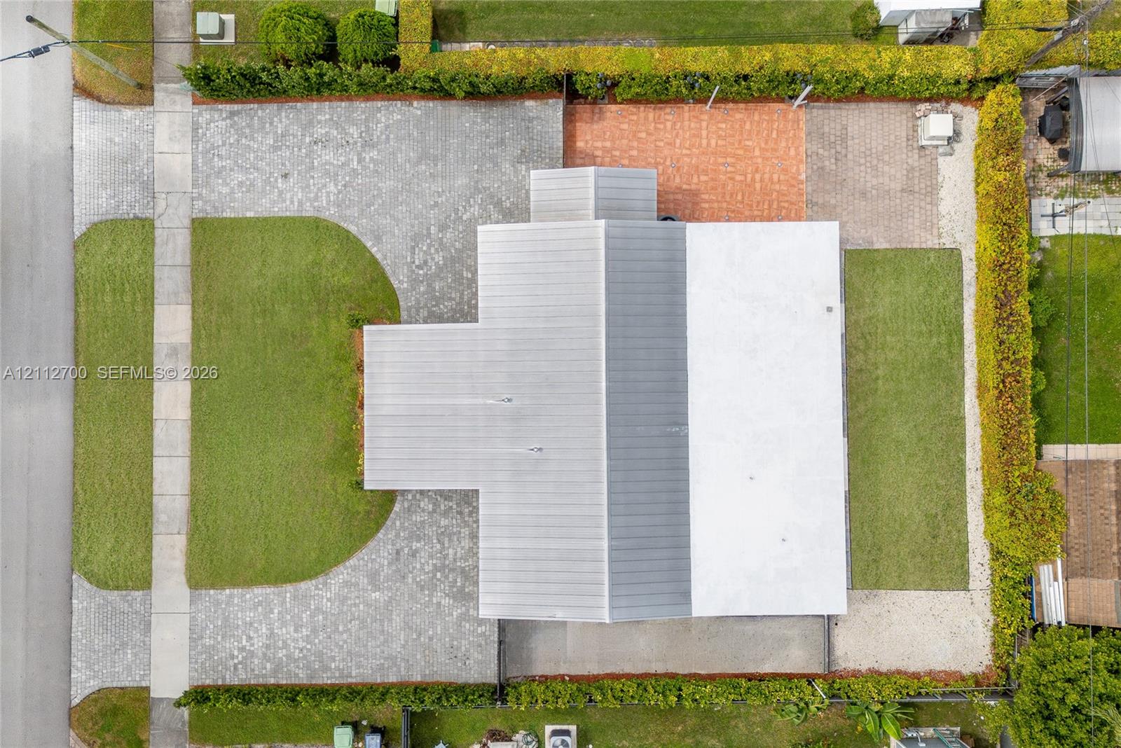 Aerial view featuring a metal roof, expansive paved driveway, dedicated brick-paver patio, manicured lawn areas, and perimeter privacy hedges