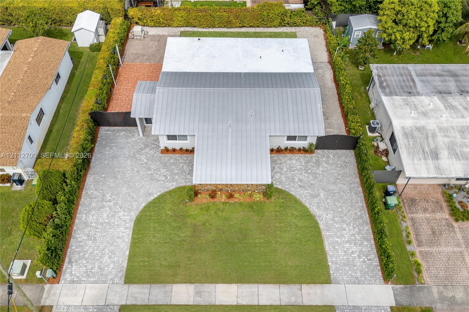 Aerial view featuring a metal roof, expansive paver driveway, manicured lawn, and perimeter privacy hedges