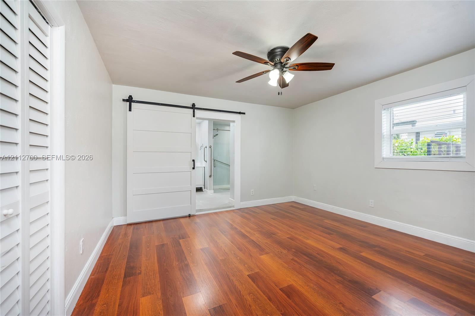 Room featuring wood-finish flooring, a white sliding barn-style door, ceiling fan with light fixture, louvered closet doors, and window with blinds