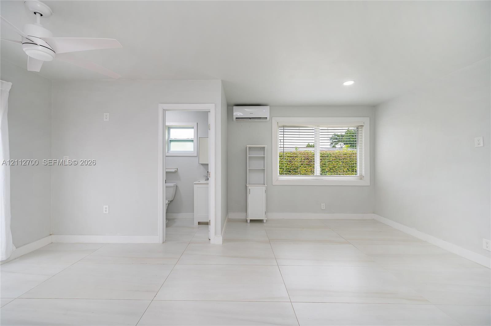 Large-format tile flooring, ceiling fan with integrated lighting, wall-mounted ductless air conditioning unit, and expansive window with horizontal blinds