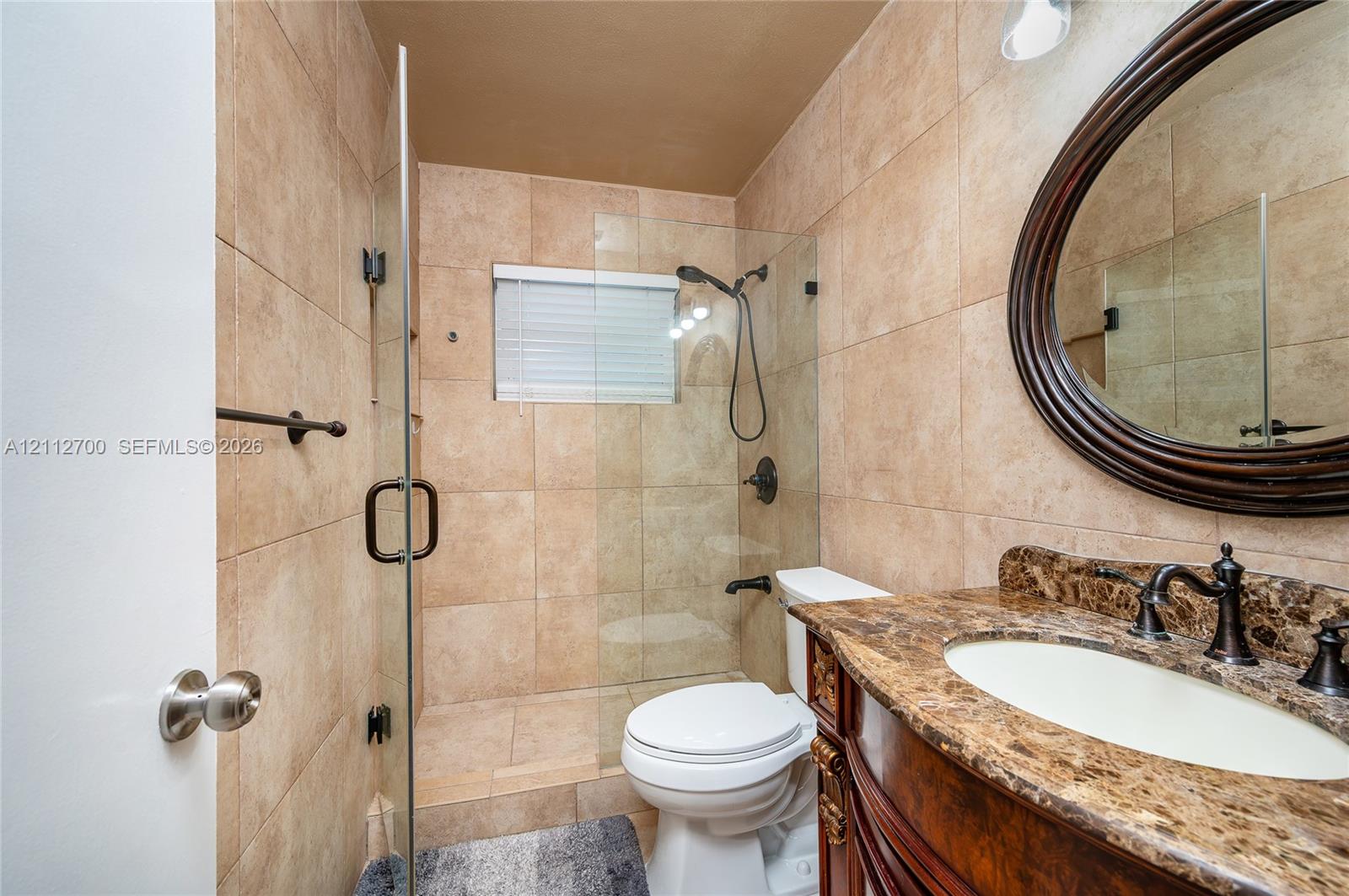 Bathroom featuring tiled walls, a glass-enclosed shower with a handheld sprayer, an oval vanity mirror with a dark wood frame, a stone-surface countertop, and a recessed sink with dark metal fixtures