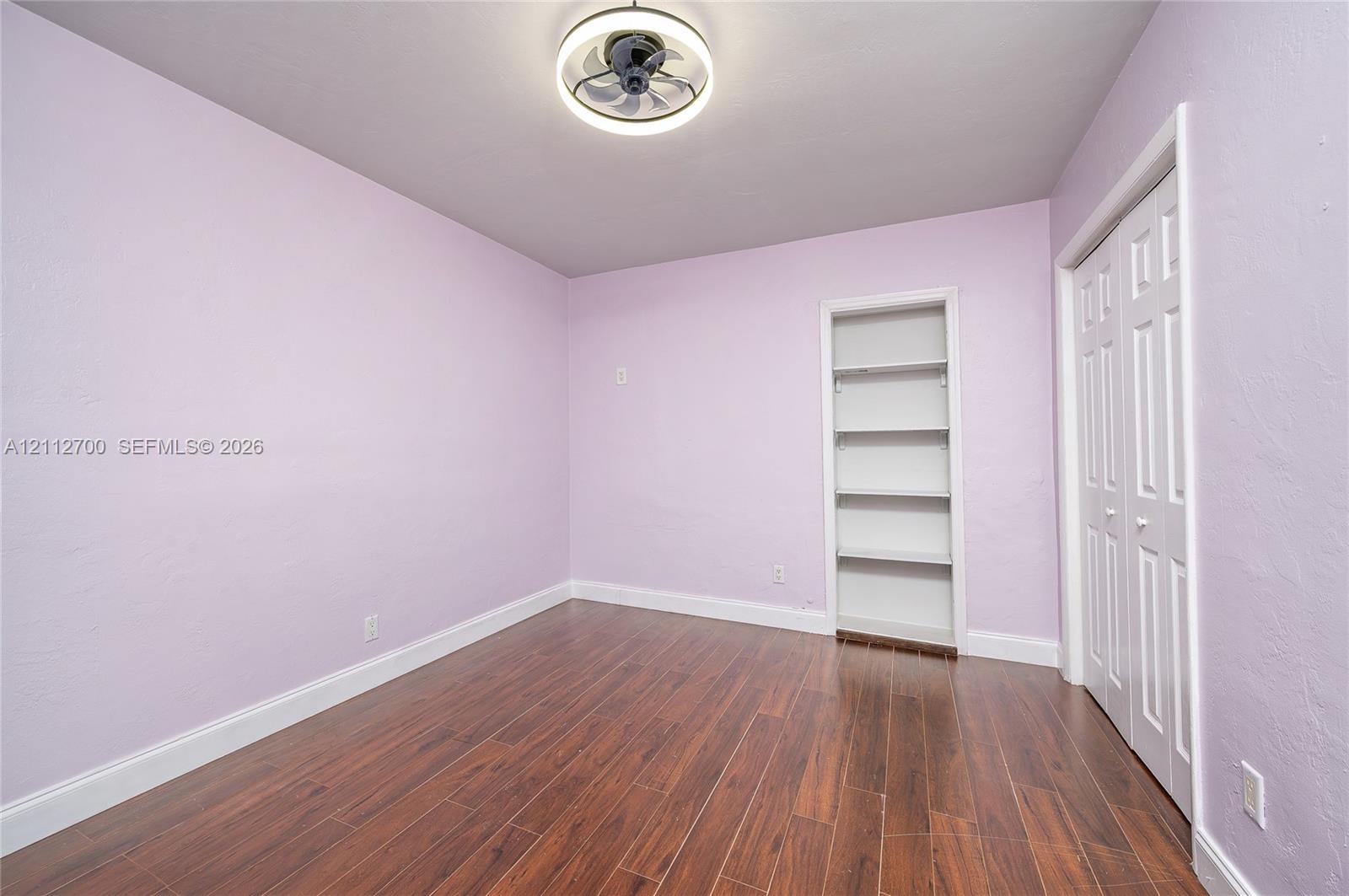 Interior space featuring wood-finish flooring, a built-in recessed wall shelving unit, double-door closet, and modern circular ceiling fan with integrated lighting