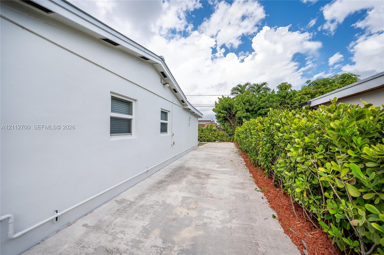 Side exterior featuring smooth stucco siding, concrete paved pathway, and mature hedgerow landscaping with organic mulch