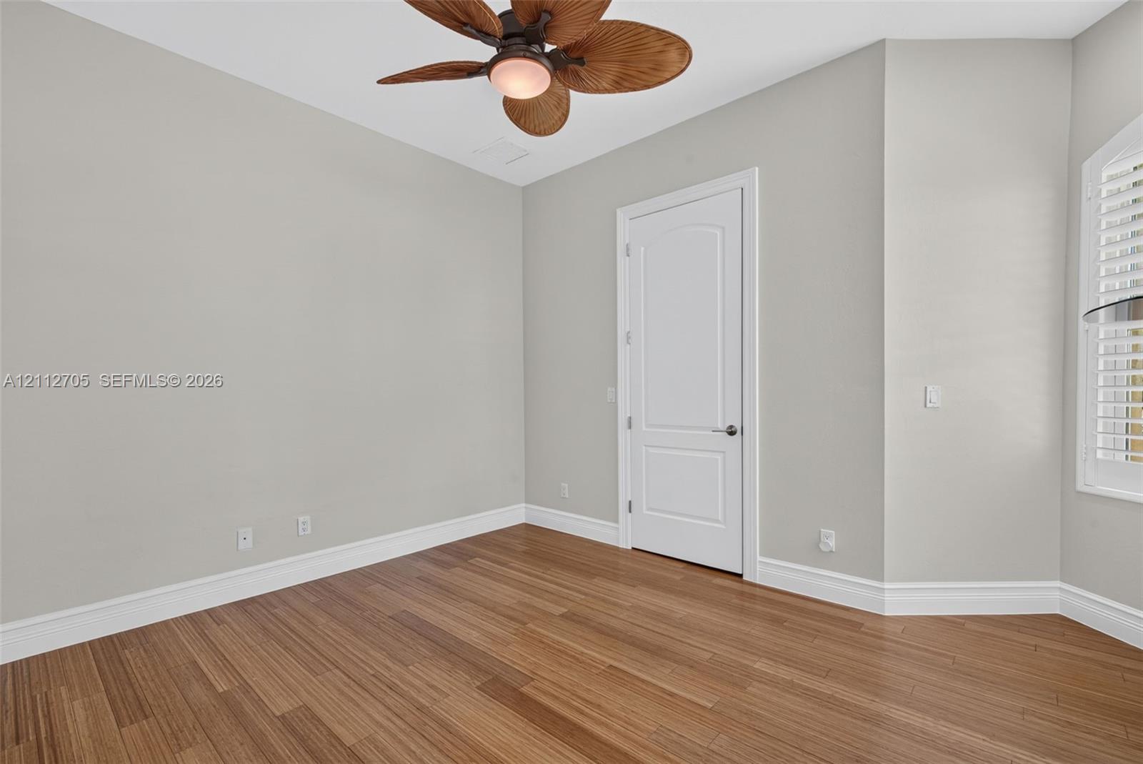 Room featuring wood-finish flooring, a neutral wall palette, a multi-blade ceiling fan with integrated lighting, a single-panel arch-top interior door, and a multi-pane window with white shuttered blinds