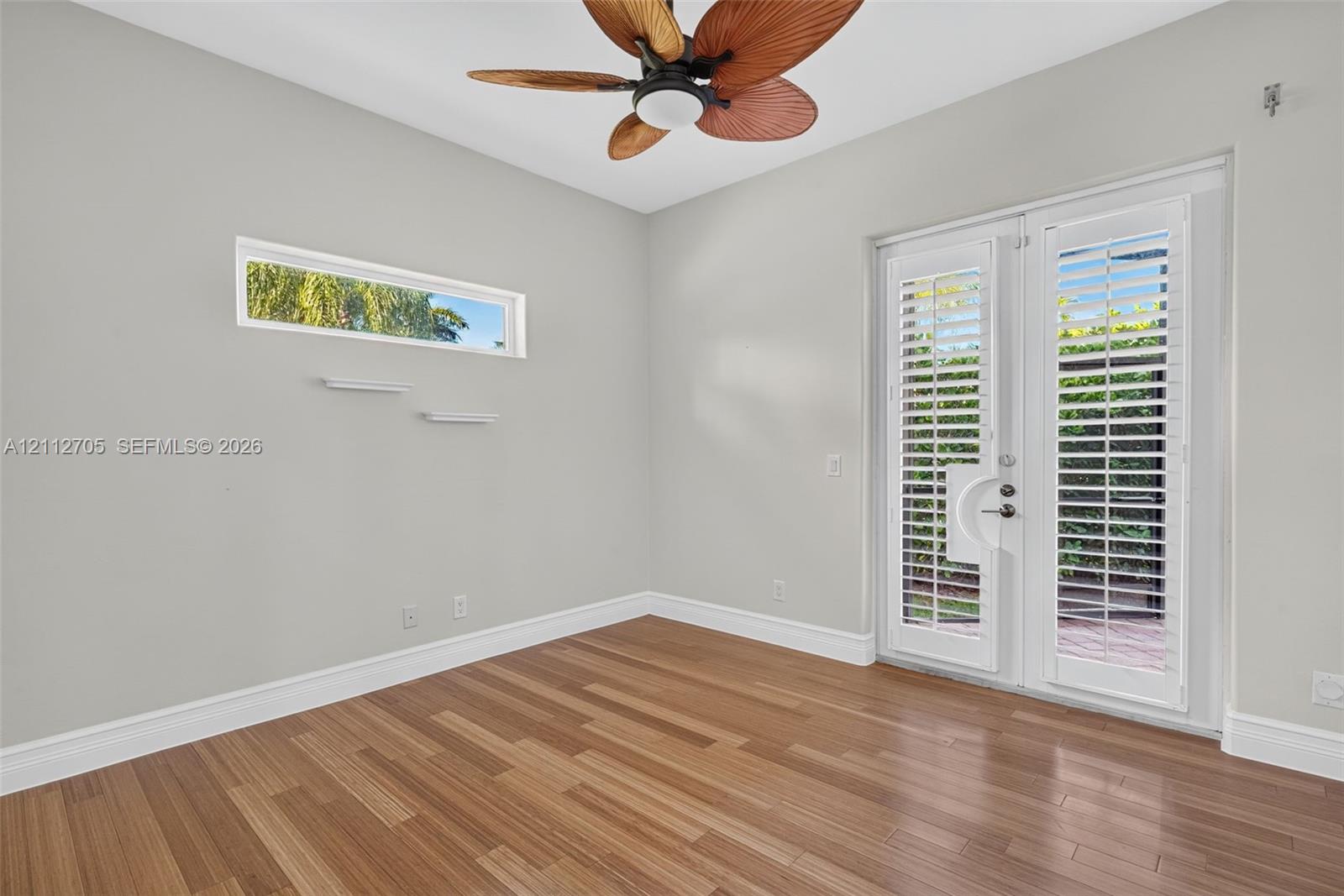 Room featuring wood-finish flooring, a horizontal transom window, French doors with integrated shutter blinds, and a ceiling fan with palm-leaf style blades