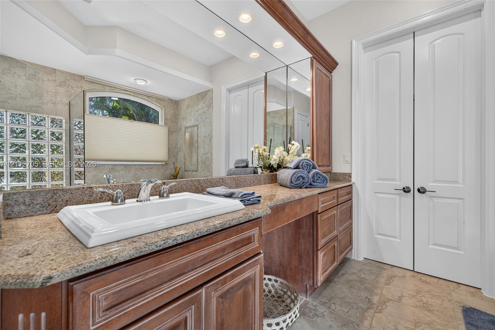 Custom wooden cabinetry featuring a dual-basin vanity, stone countertop, integrated full-width mirror, and glass block accent window