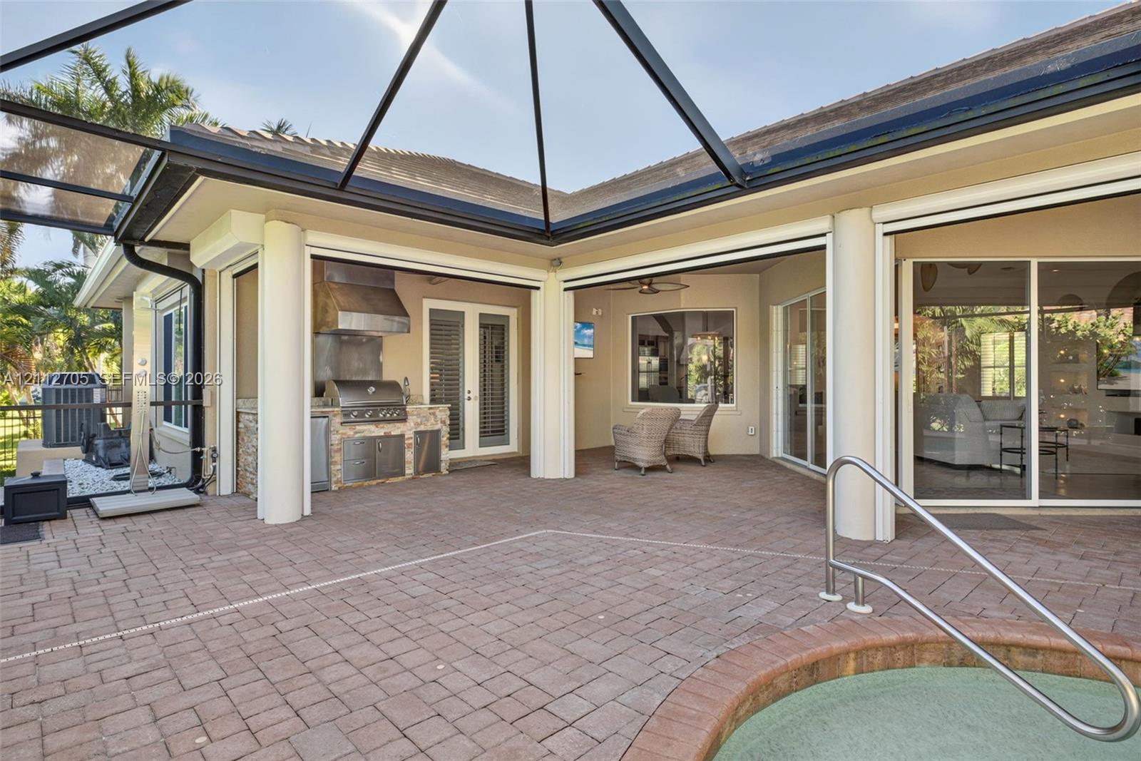 Covered lanai featuring a brick-paver deck, built-in outdoor kitchen with stainless steel grill and hood, and an integrated spa