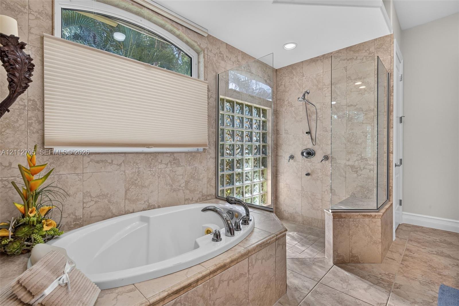 Soaking tub with deck-mounted faucet, walk-in glass-enclosed shower, natural stone-finish wall and floor tile, arched window, and glass block accent window
