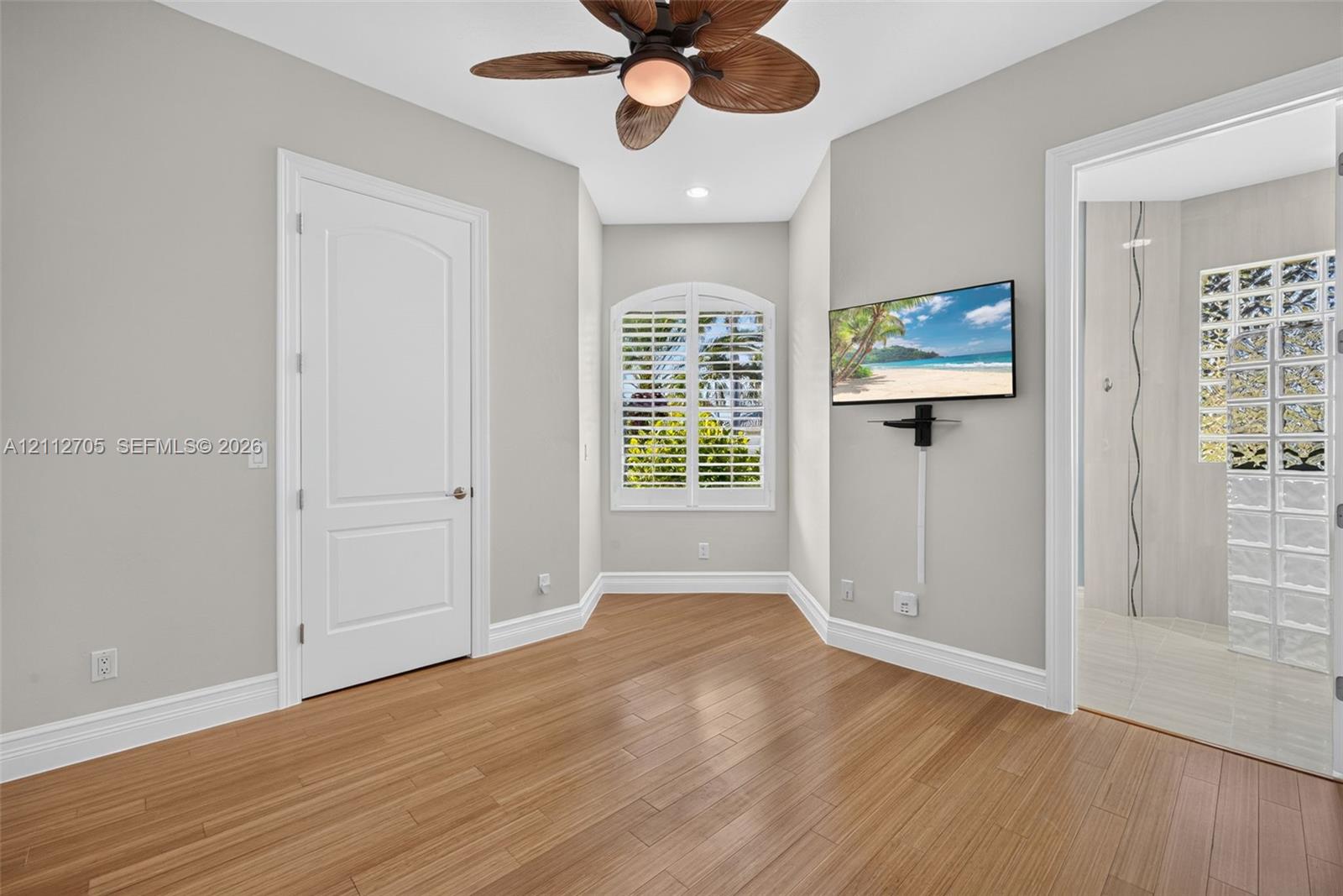 Room featuring wood-finish flooring, a ceiling fan with palm-leaf blades, arched window with plantation shutters, and an entrance to a bathroom with a glass block wall
