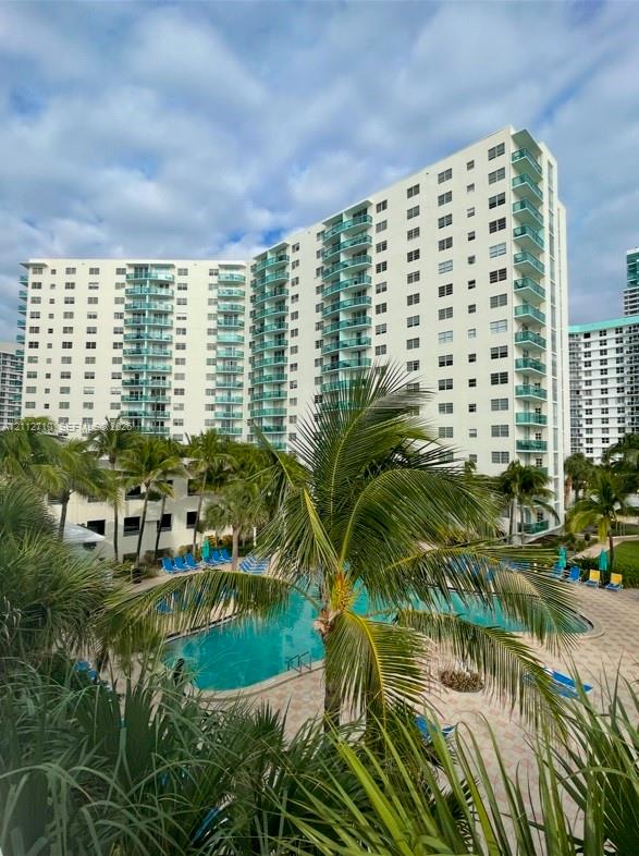 Multi-story high-rise building featuring private balconies with glass railings, surrounding swimming pool, stone-paver pool deck, and mature palm landscaping
