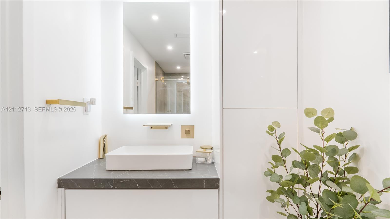 Modern vanity featuring a rectangular vessel sink, dark stone-finish countertop, integrated tall cabinetry, gold-tone hardware, and an LED-backlit mirror