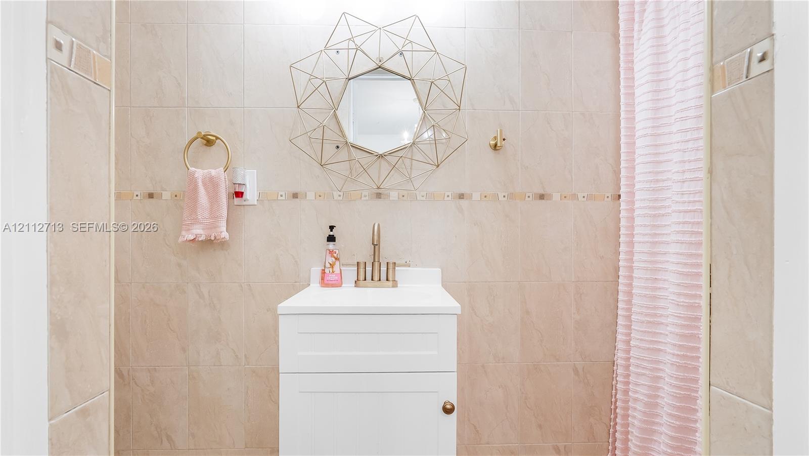 White vanity with a rectangular sink, metallic faucet, wall-mounted circular geometric mirror, and floor-to-ceiling tile featuring a decorative accent border