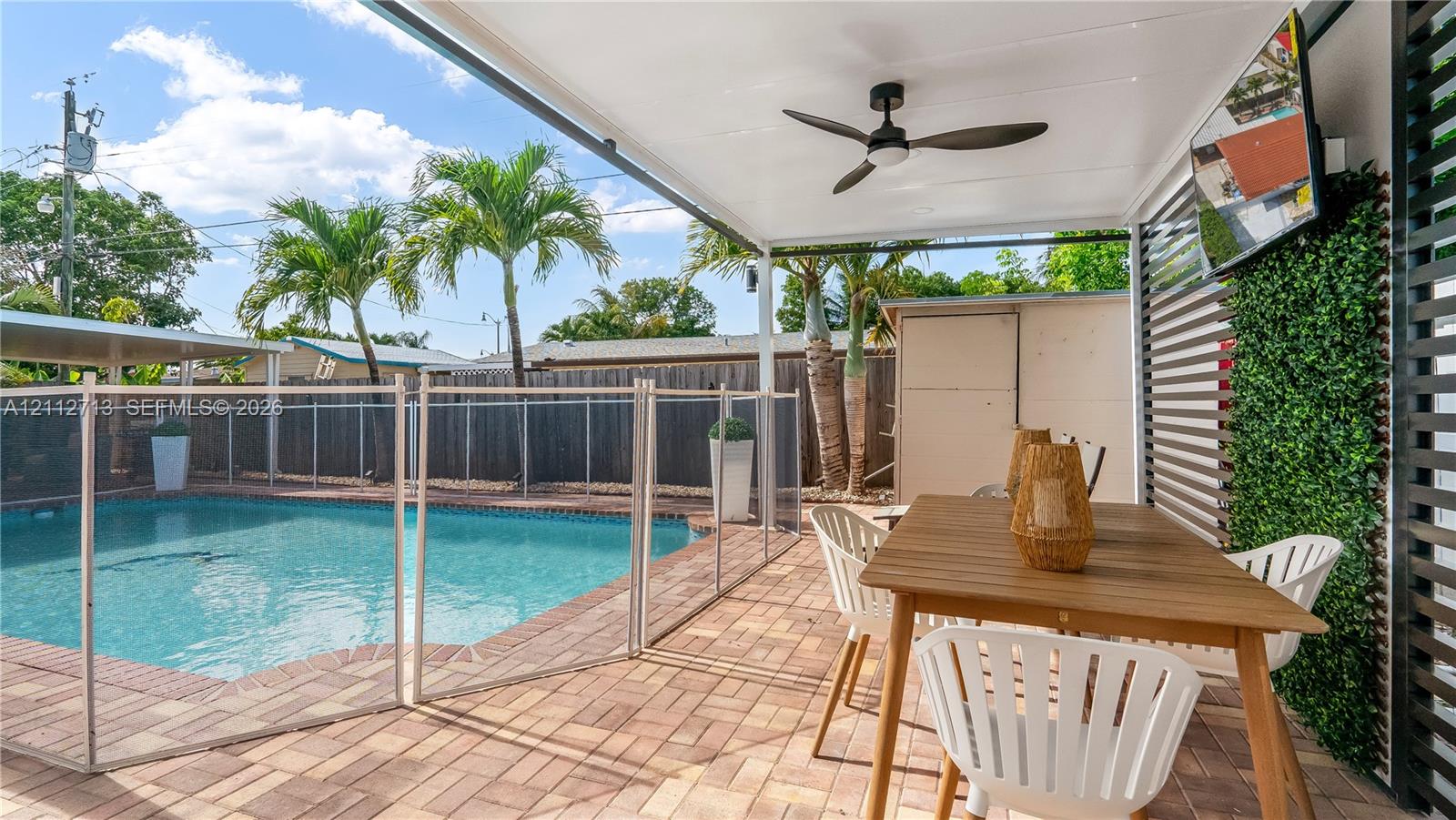 Covered patio featuring a ceiling fan, paver flooring, horizontal slat wall, and attached vertical garden wall, leading to a swimming pool with a safety enclosure Covered patio featuring a ceiling fan, paver flooring, horizontal slat wall, and attached vertical garden wall, leading to a swimming pool with a safety enclosure