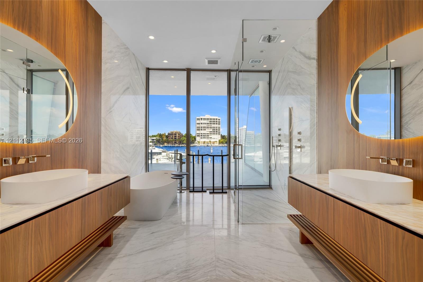 Dual floating wood-finish vanities with vessel sinks and integrated backlit mirrors, freestanding soaking tub, expansive stone-tiled floor, and a glass-enclosed walk-in shower