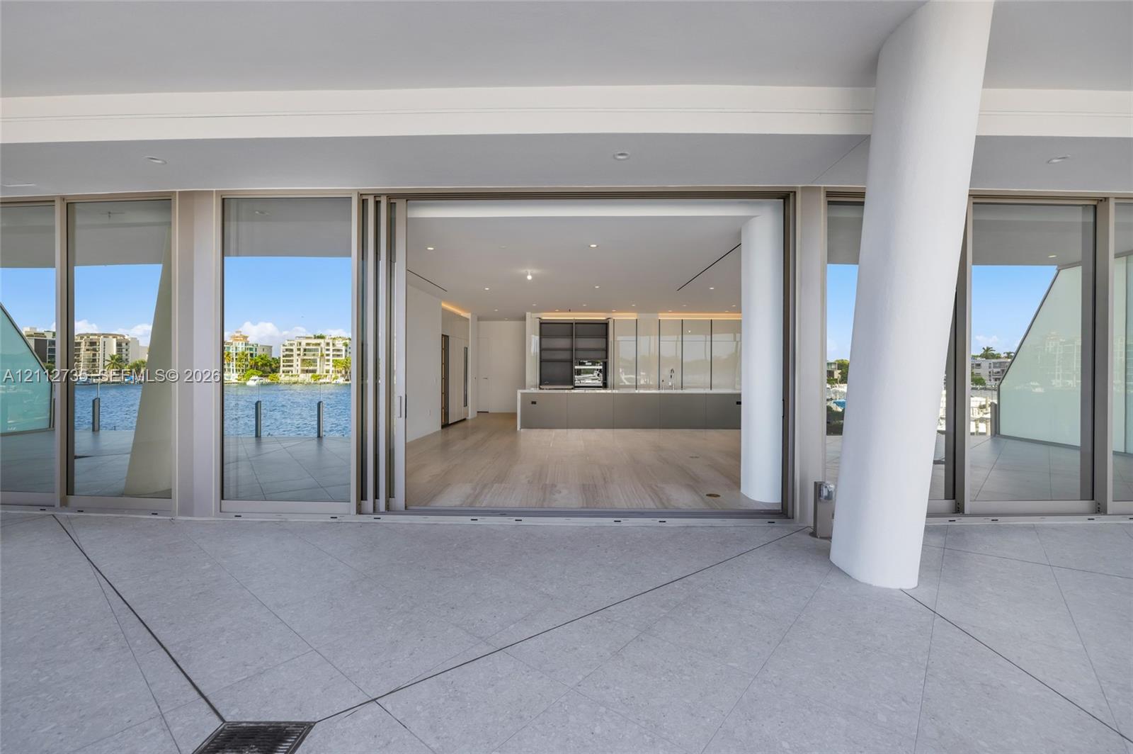 Covered terrace featuring stone tile flooring and a structural cylindrical column, spanning wide sliding glass doors that reveal wood-finish flooring and built-in kitchen cabinetry
