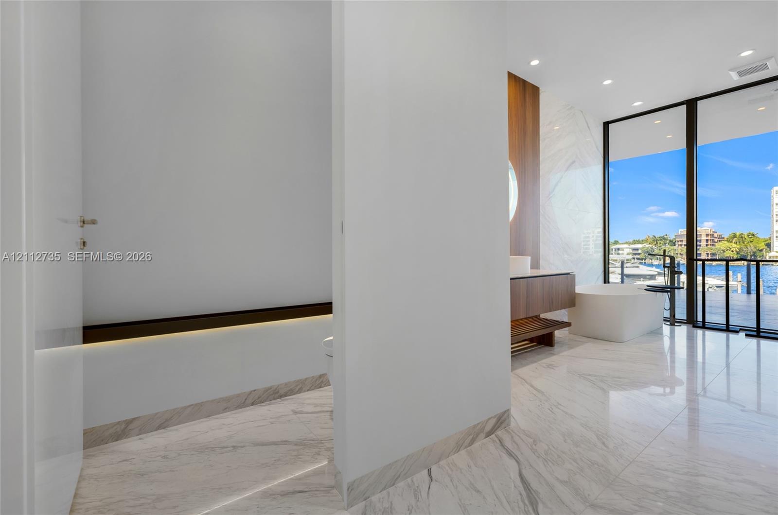 Stone-tiled flooring, wall-mounted wood-finish vanity, freestanding soaking tub, recessed ceiling lighting, and floor-to-ceiling glass sliding doors
