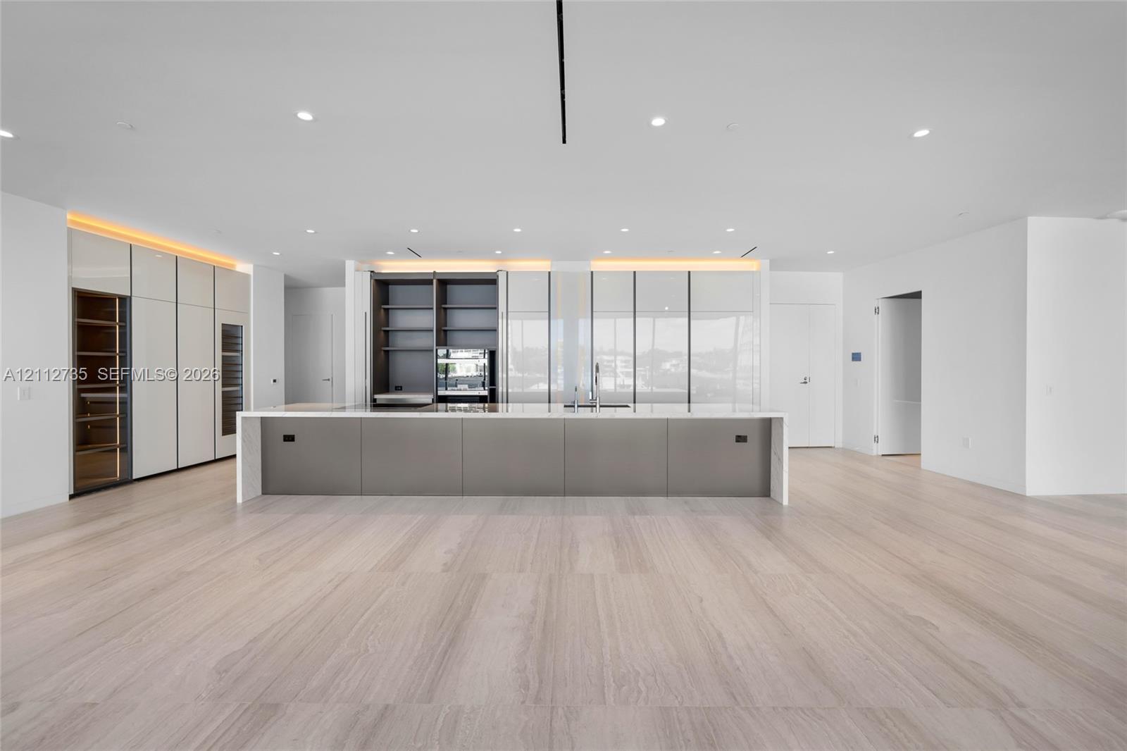 Minimalist kitchen featuring an expansive center island with a stone countertop, floor-to-ceiling cabinetry with integrated display shelving, built-in wine refrigeration, and light-toned wood-finish flooring