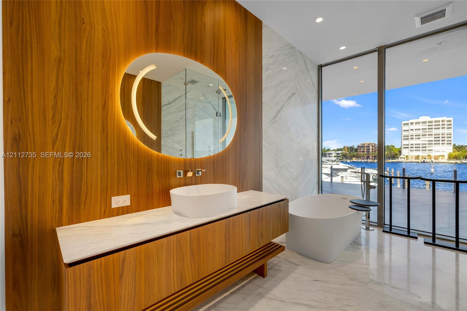 Floating wood-finish vanity with an integrated countertop, oval back-lit mirror, vessel sink, freestanding soaking tub, and floor-to-ceiling glass paneling