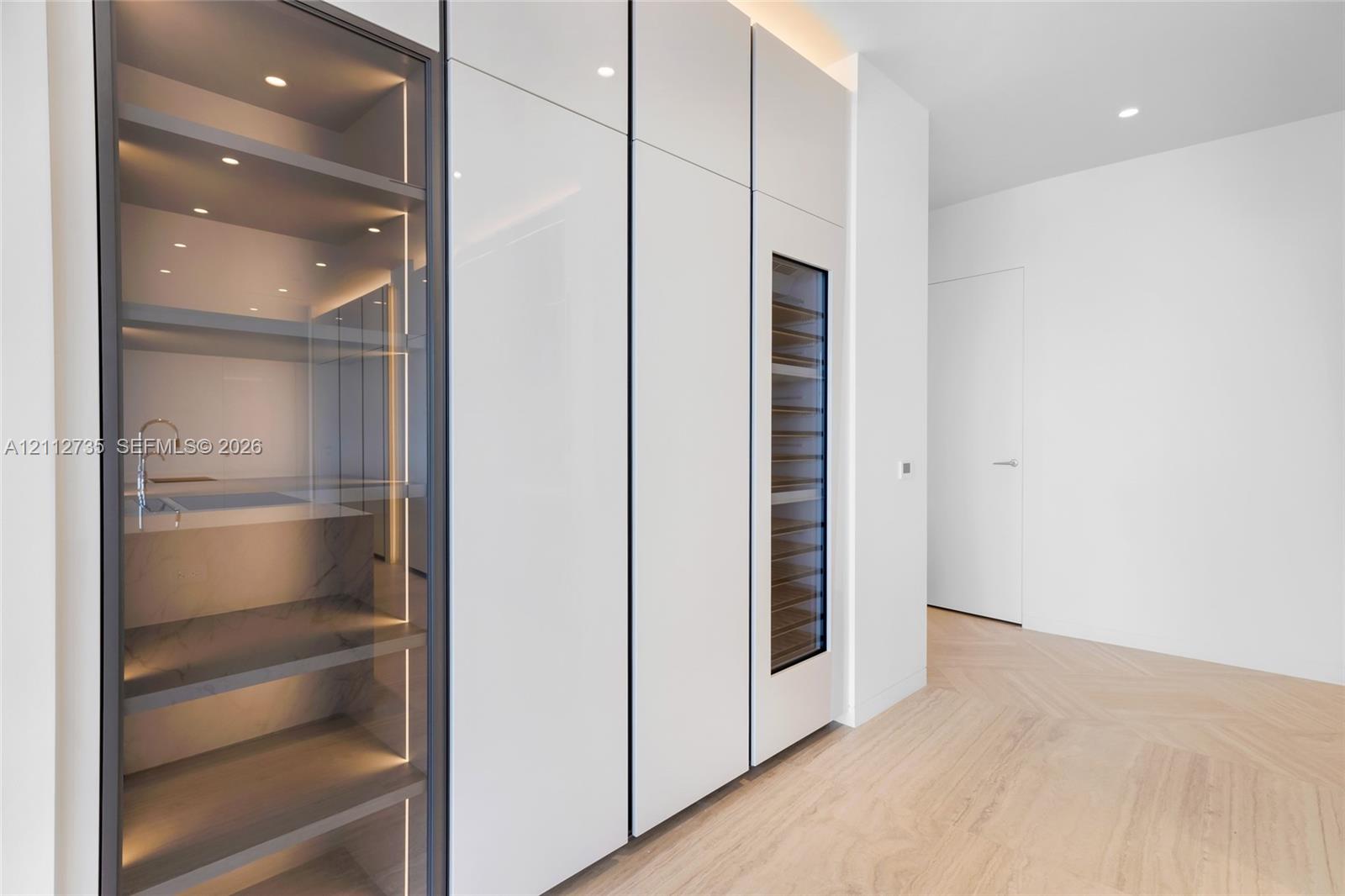 Custom millwork featuring floor-to-ceiling high-gloss cabinetry, integrated wine refrigeration, glass-front display shelving with LED illumination, and chevron wood-finish flooring