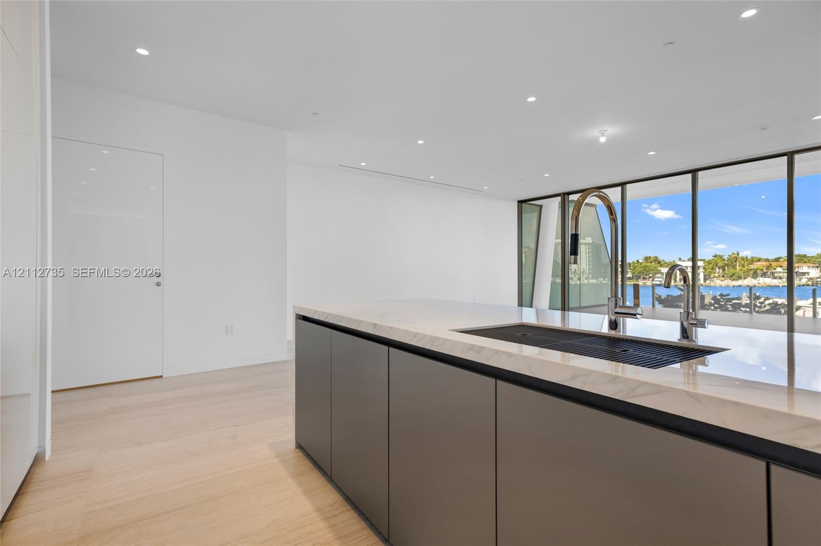 Kitchen island featuring flat-panel cabinetry, light-toned stone-finish countertop, integrated sink, and polished metal faucet, complemented by wood-finish flooring and floor-to-ceiling windows
