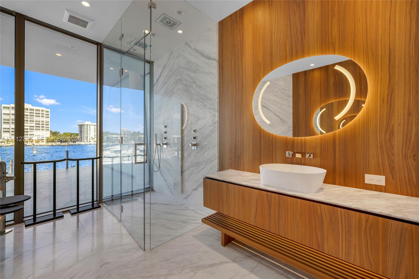 Floating wood-finish vanity with a stone countertop and vessel sink, illuminated oval mirror, walk-in glass-enclosed shower, stone-surround shower walls, and full-height floor-to-ceiling windows