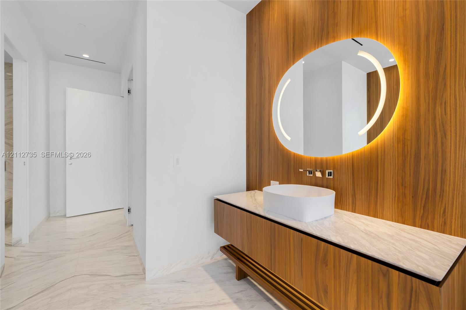 Floating wood-finish vanity with a stone countertop, white vessel sink, wall-mounted faucet, and custom wood-paneled wall featuring an integrated circular mirror