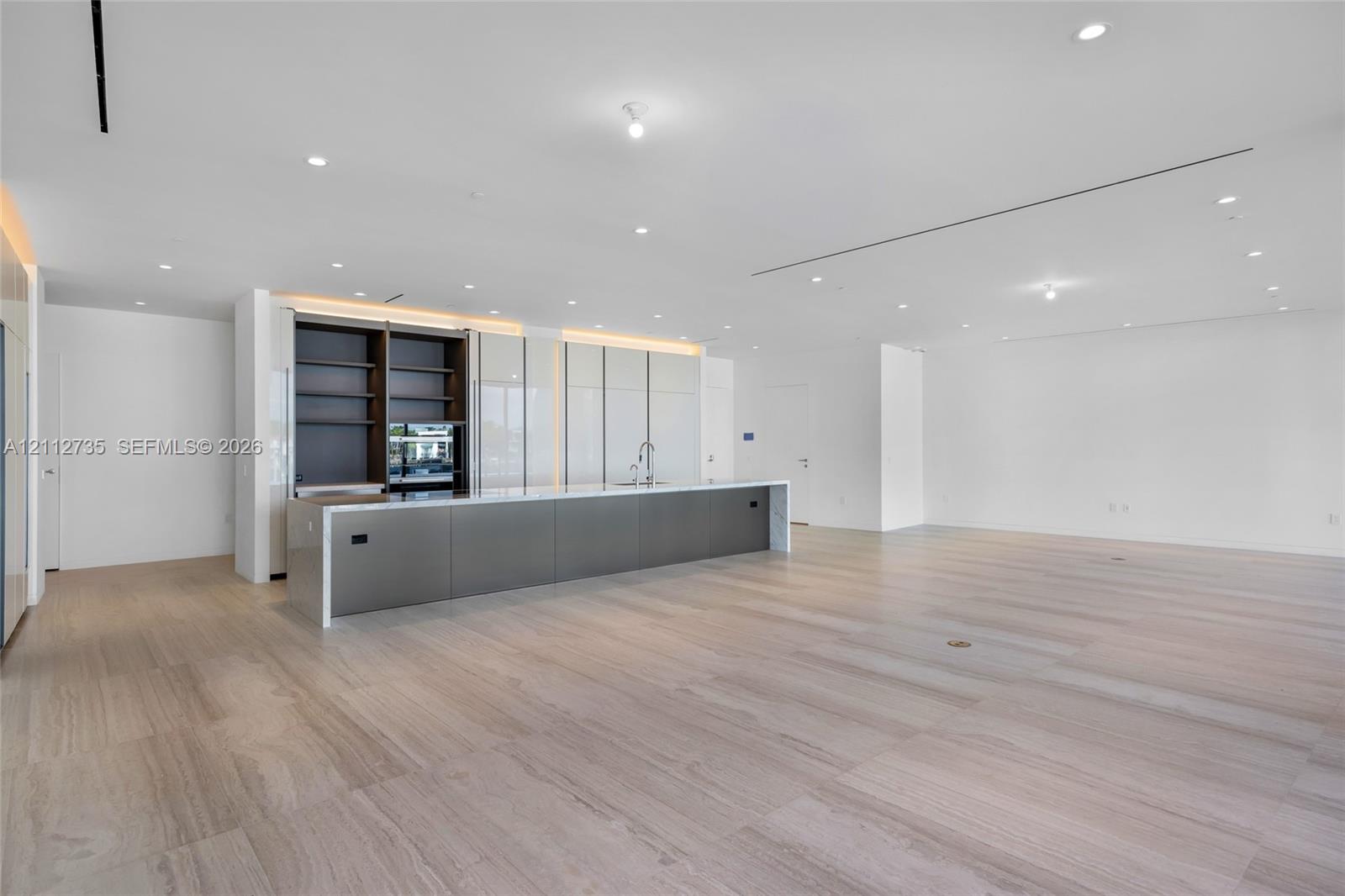 Minimalist kitchen featuring an expansive island with metallic cabinetry, light-toned stone countertops, built-in shelving, integrated lighting, and wood-finish flooring