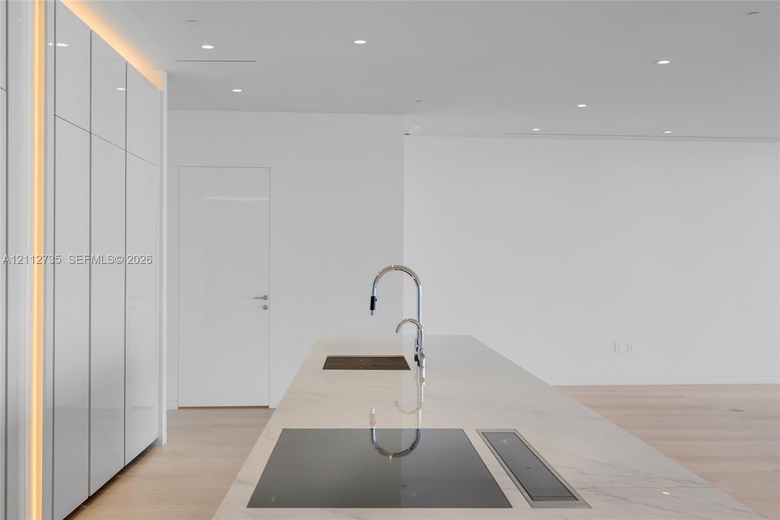 Minimalist kitchen island featuring a stone countertop, flush-mount cooktop, integrated downdraft ventilation, and a high-arc faucet, complemented by floor-to-ceiling cabinetry with integrated LED lighting and light-toned wood-finish flooring