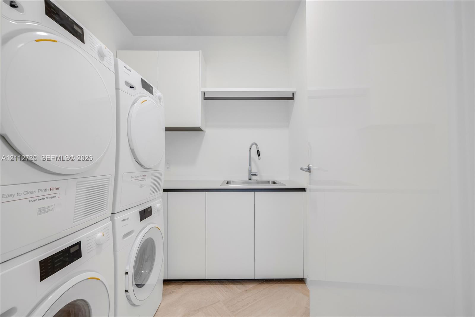 Utility area featuring stacked laundry appliances, white built-in cabinetry, a rectangular stainless steel sink with a high-arch pull-down faucet, and light-tone wood-finish flooring